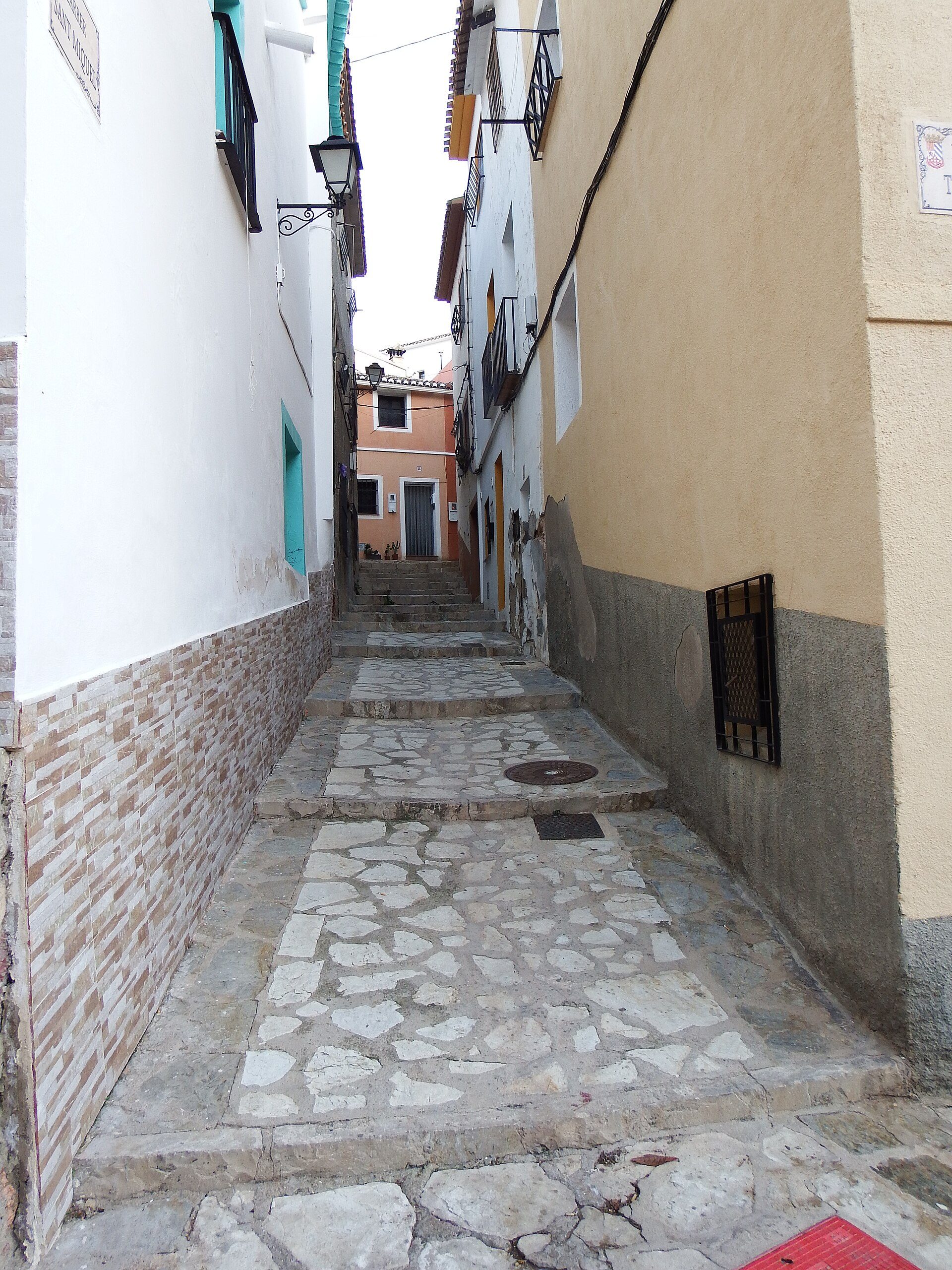 Narrow stone-paved alley with white and beige buildings, featuring balconies and a streetlamp.