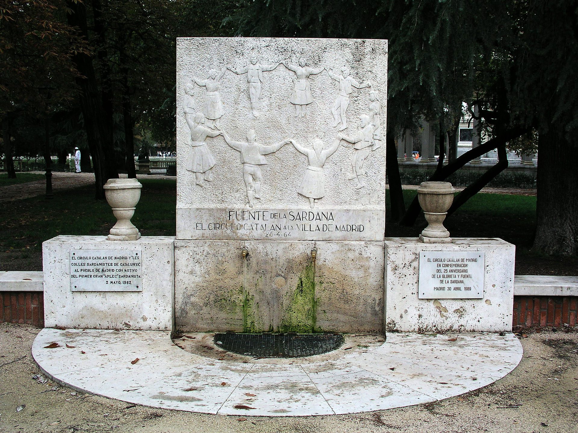 Alt text: Outdoor monument with relief sculpture, inscriptions, and two urns, surrounded by trees.