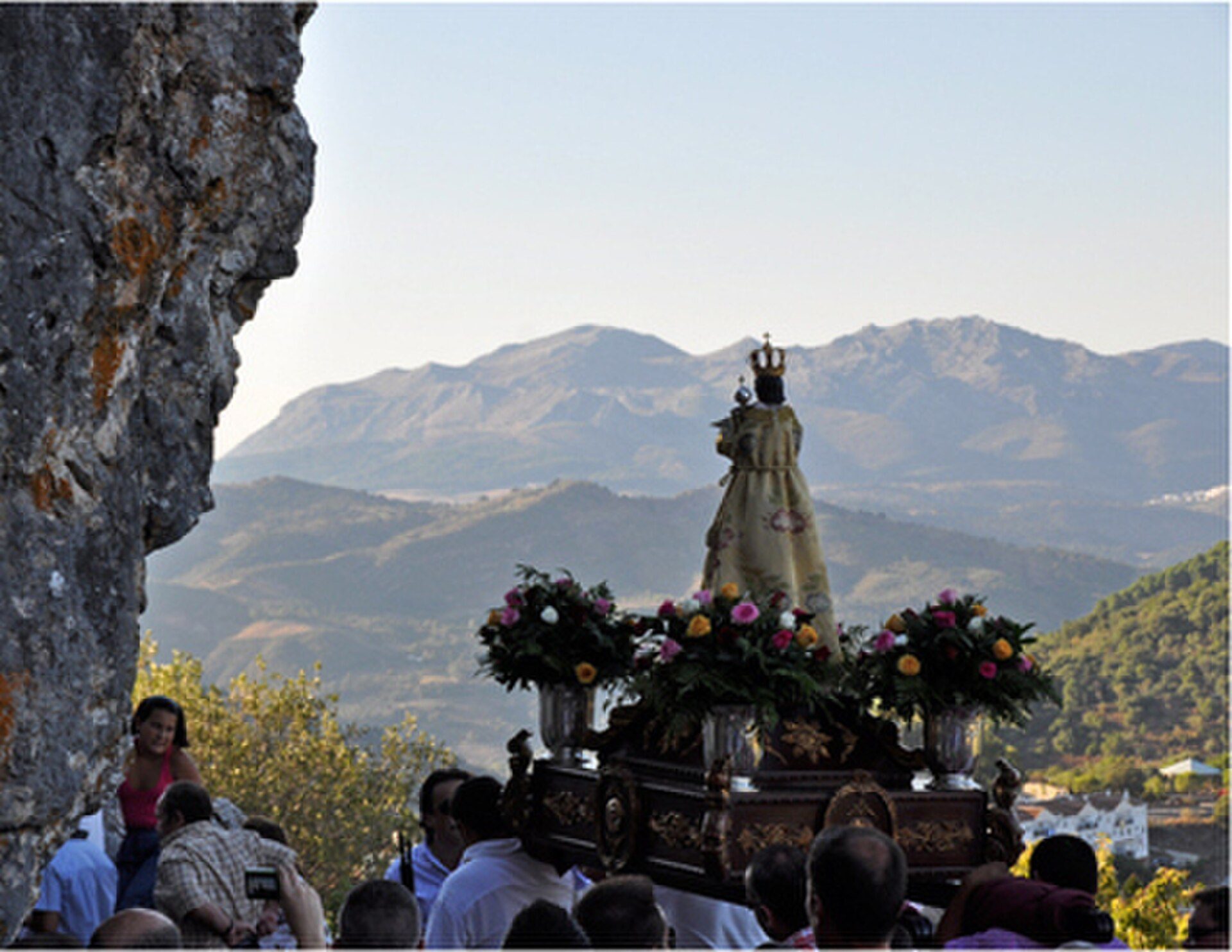 Alt text: Outdoor procession with a statue, surrounded by flowers, set against mountainous landscape.