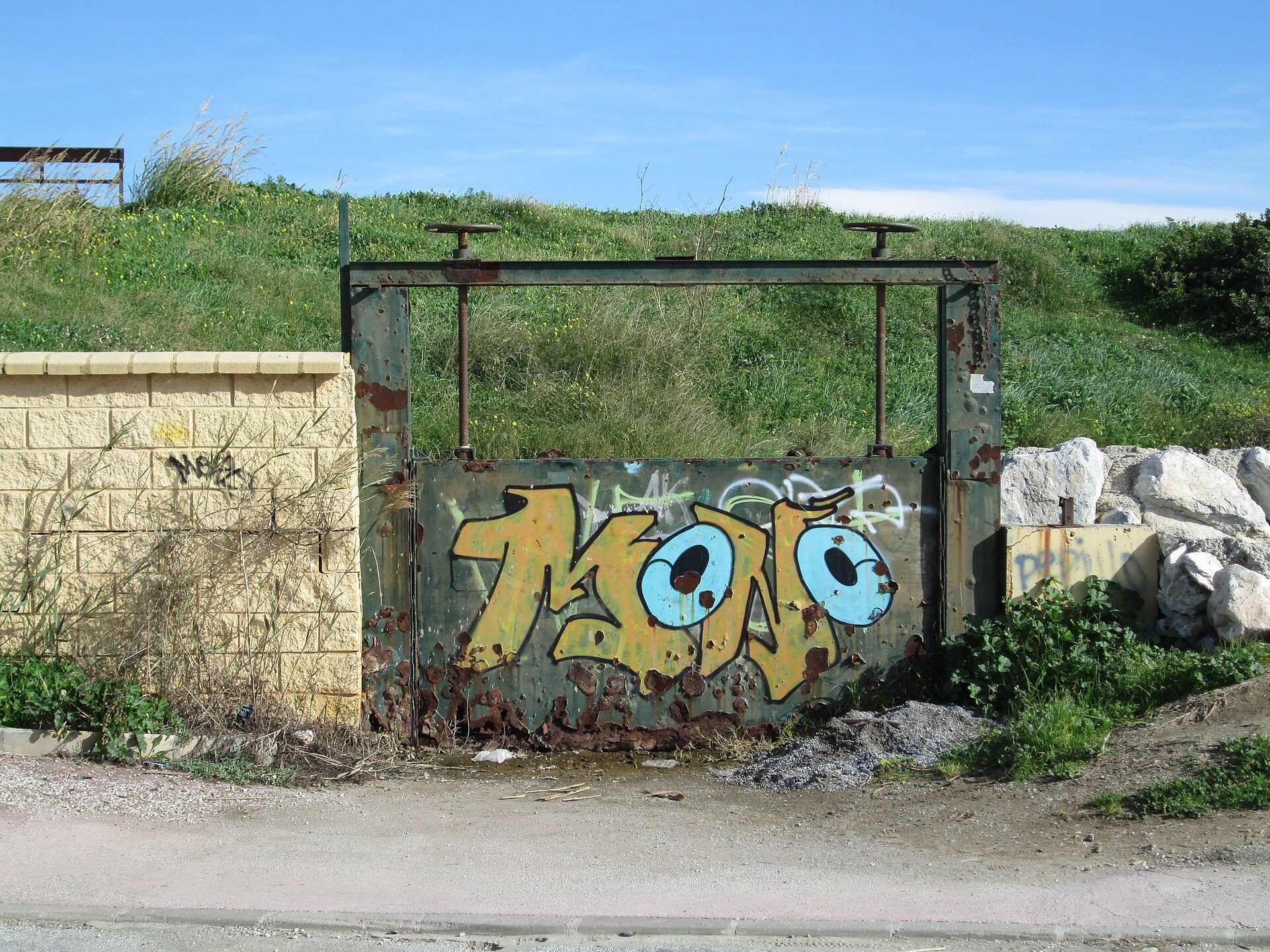 Rustic outdoor scene with graffiti on a metal gate, greenery, and a clear blue sky.