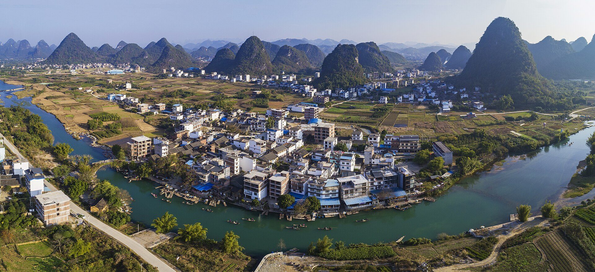 Aerial view of a riverside village with traditional houses and lush karst mountains in the background.