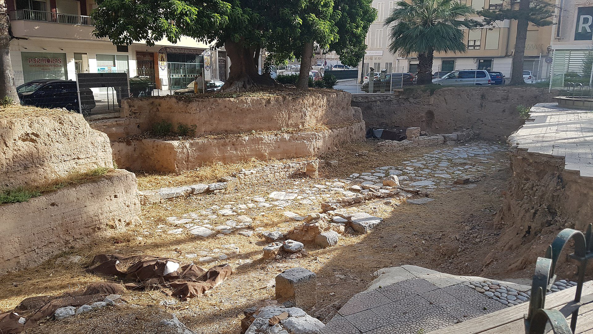 Historic site with ancient stone ruins, surrounded by modern buildings and trees.