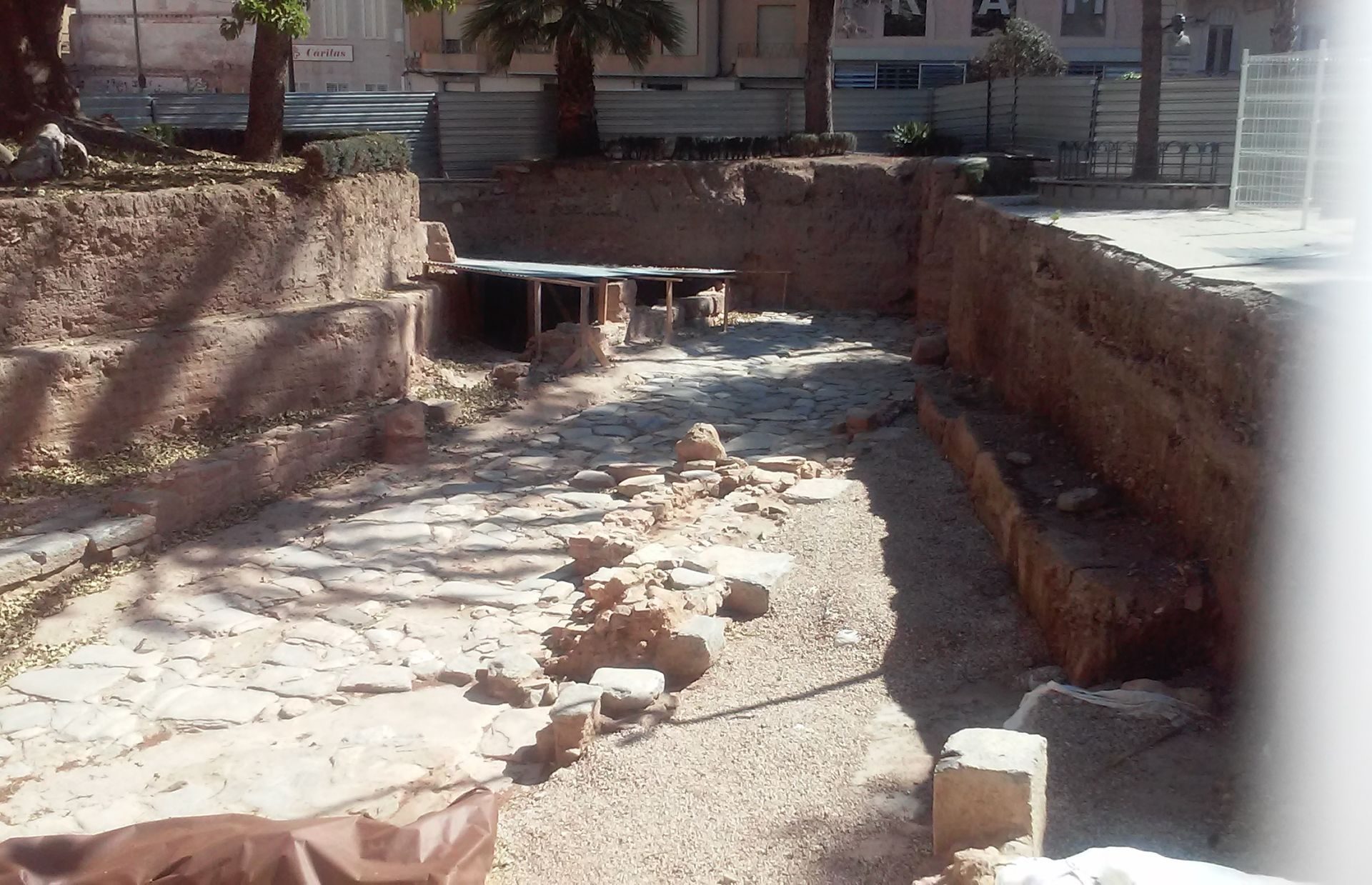Exposed stone floor and walls, outdoor courtyard with palm trees and modern building in background.