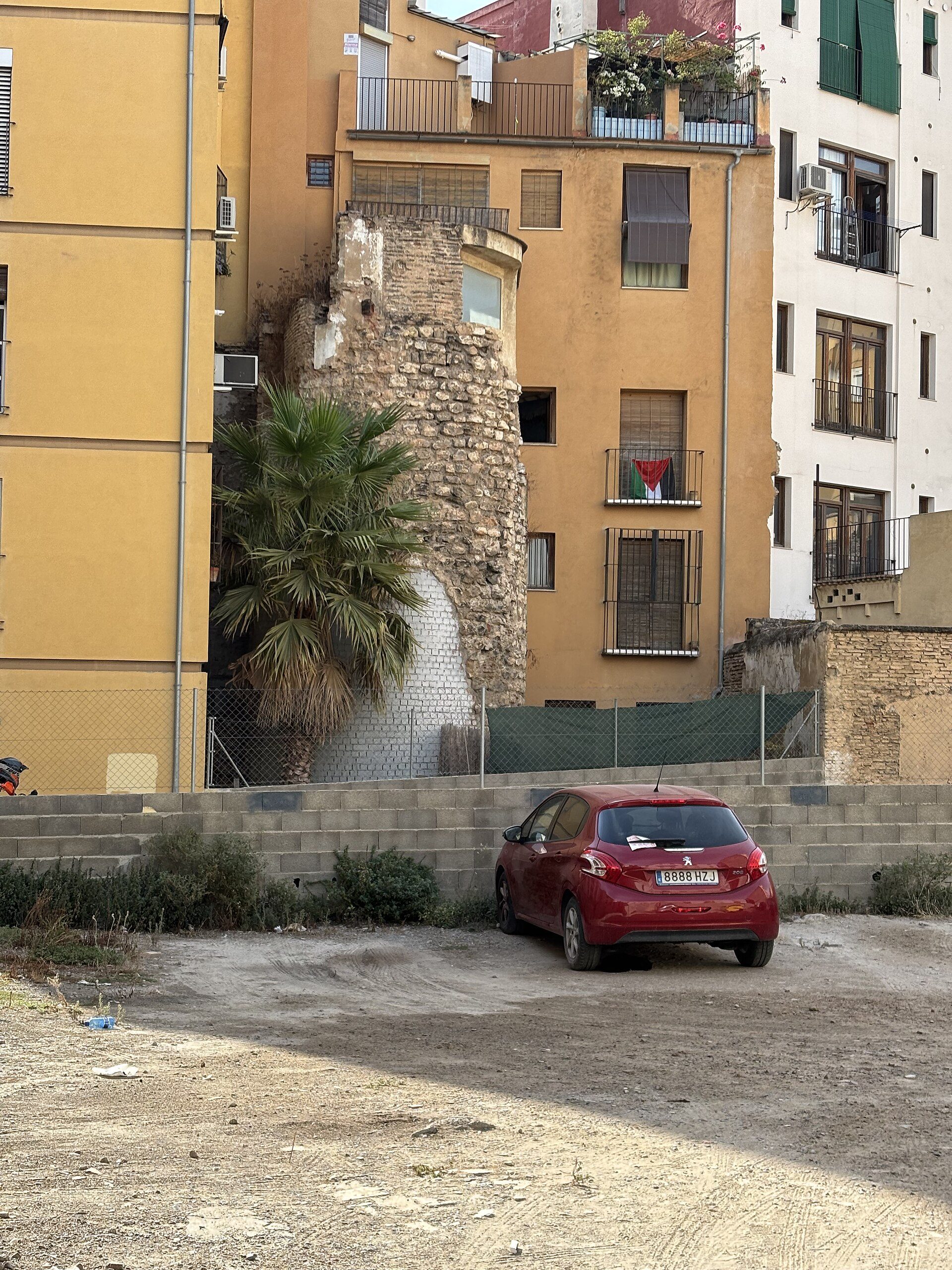 A red car parked in front of a multi-story building with balconies and a palm tree.