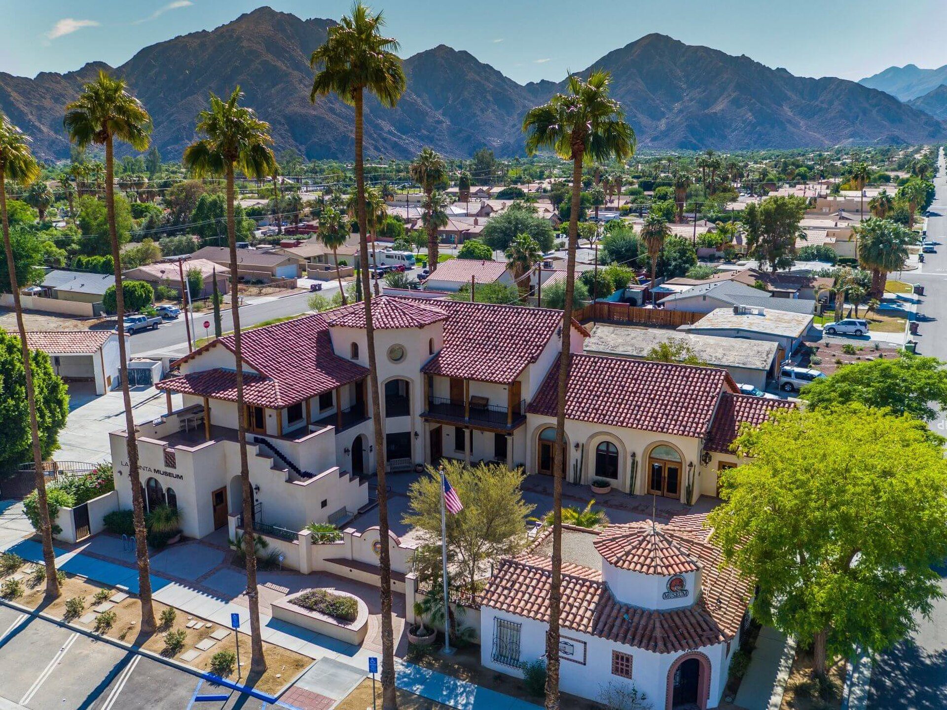 Aerial view of a two-story building with a red-tiled roof, surrounded by palm trees and mountains.