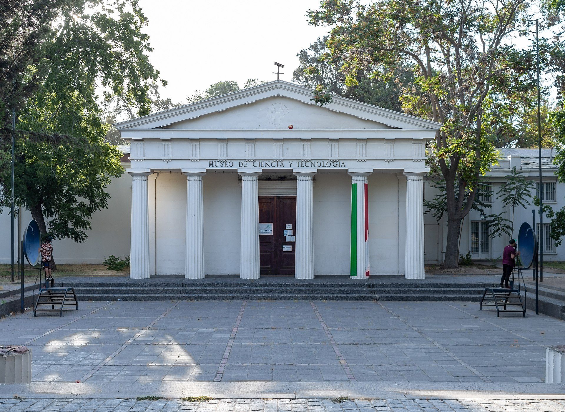Alt text: Classical building with columns, green and red flag, trees, and benches.