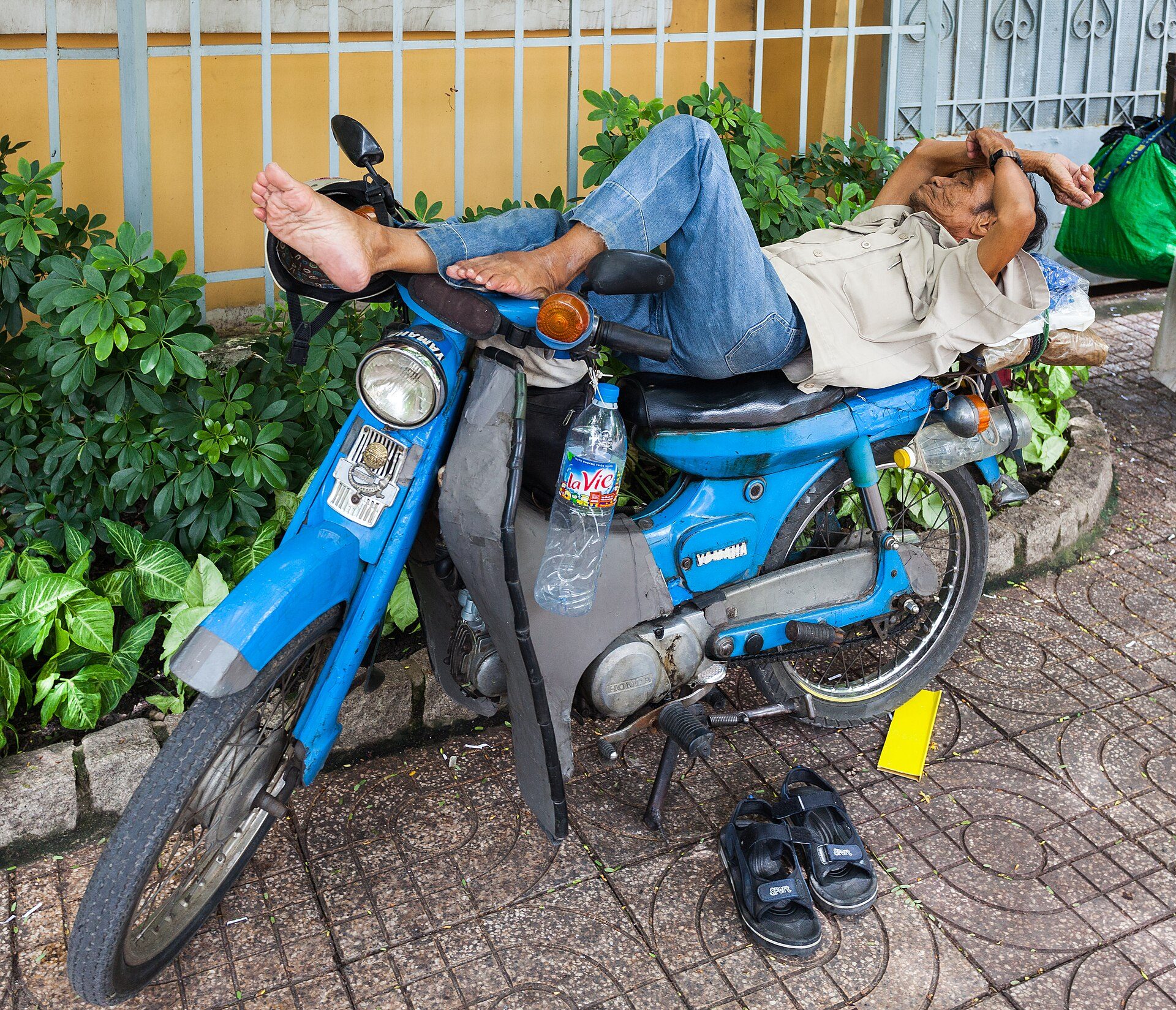 A man is lying on a blue scooter on a sidewalk, with a bottle of water and shoes nearby.