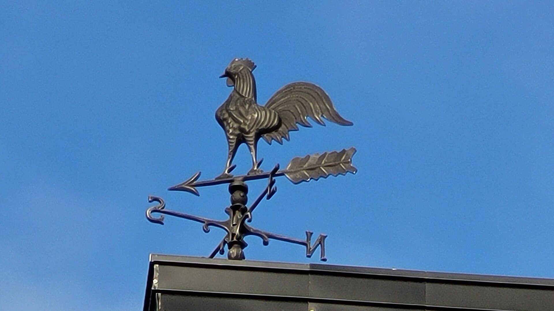 A weather vane with a rooster on top, set against a clear blue sky.