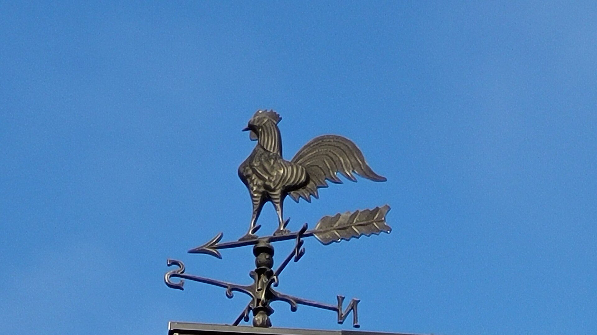 A weather vane featuring a rooster on top of a building against a clear blue sky.