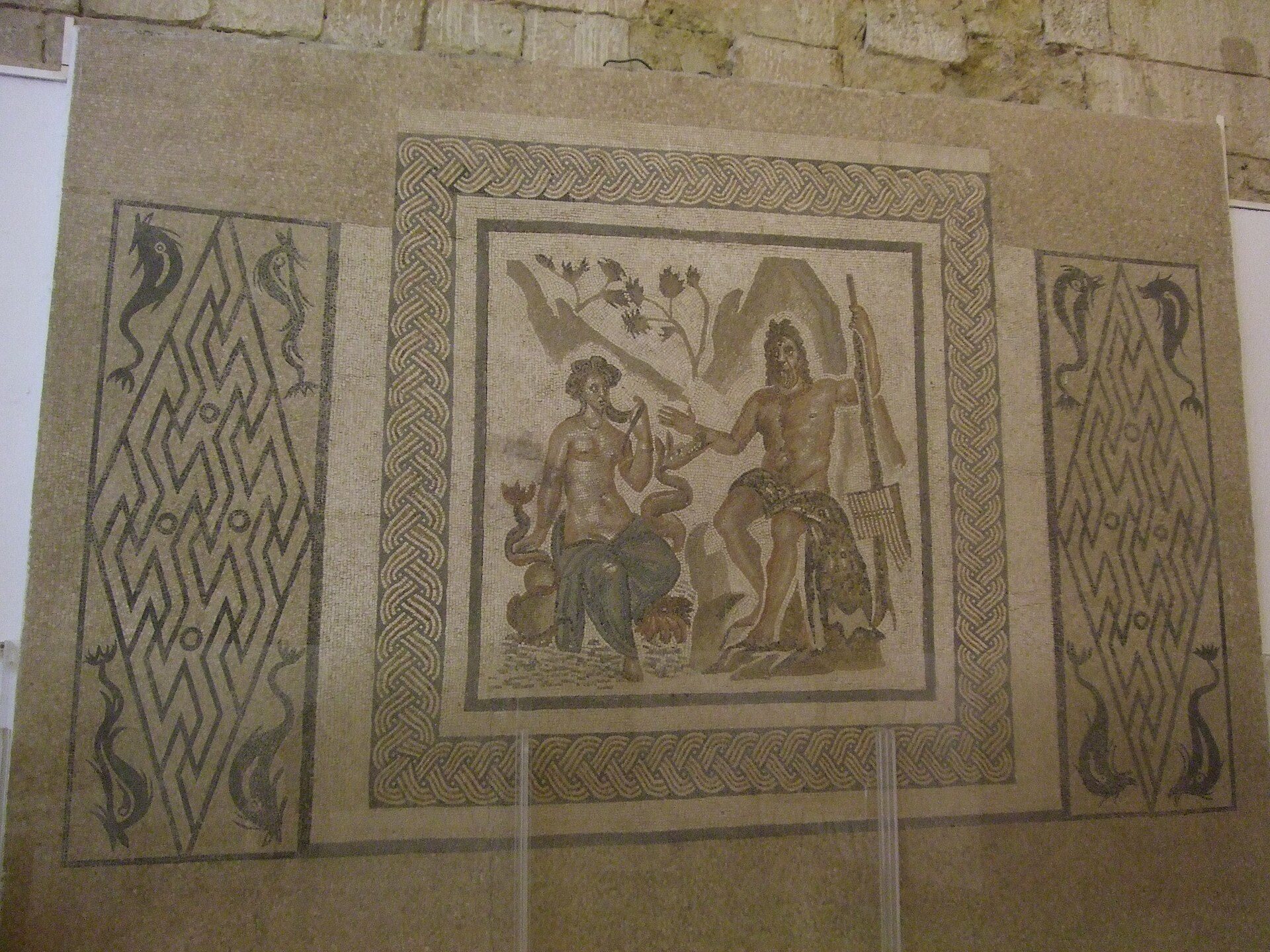 Mosaic artwork depicting two figures with decorative borders, displayed on a wall.