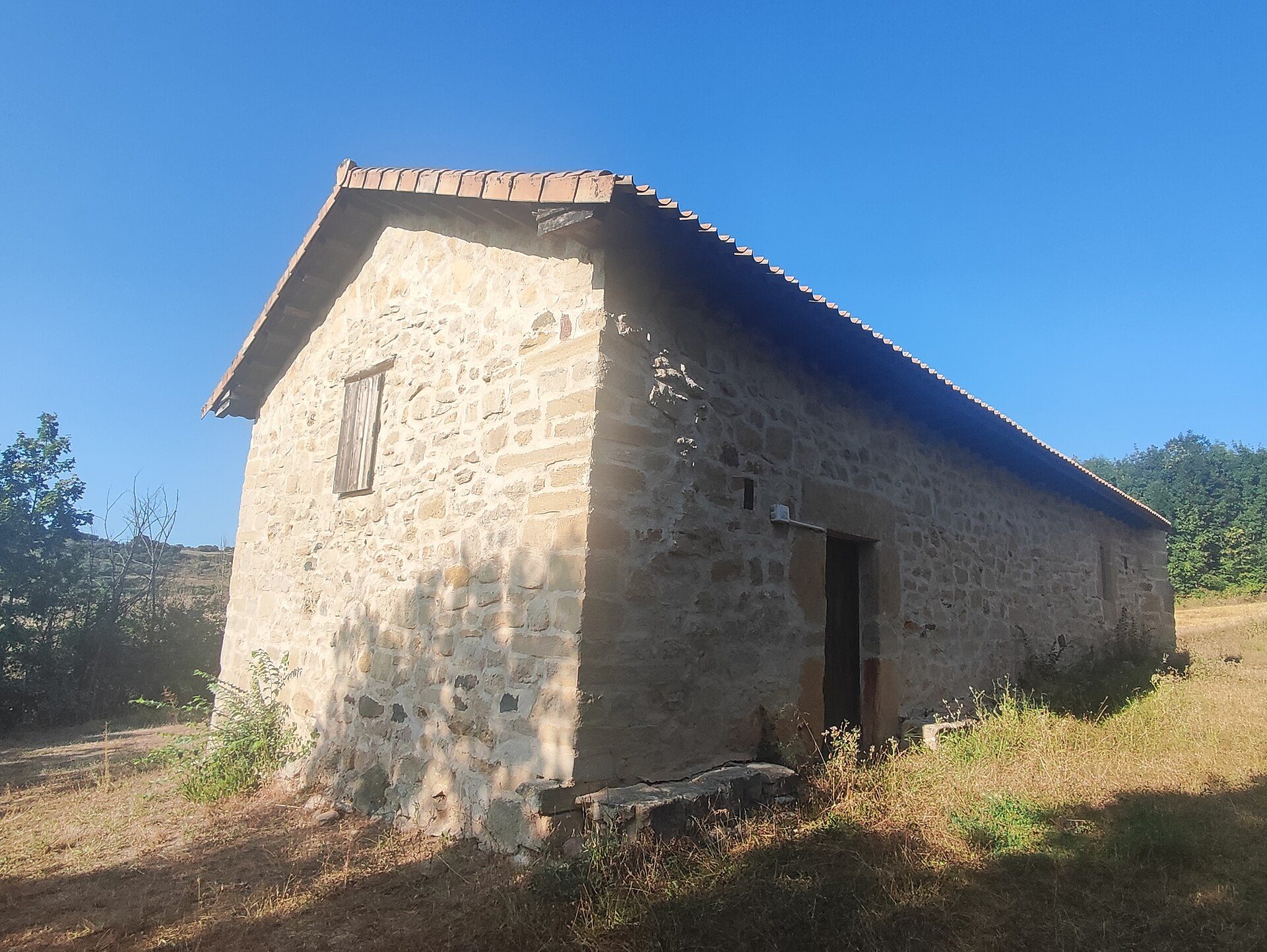 A small stone cottage with a sloped roof, featuring a single window and a door, set in a rural landscape.