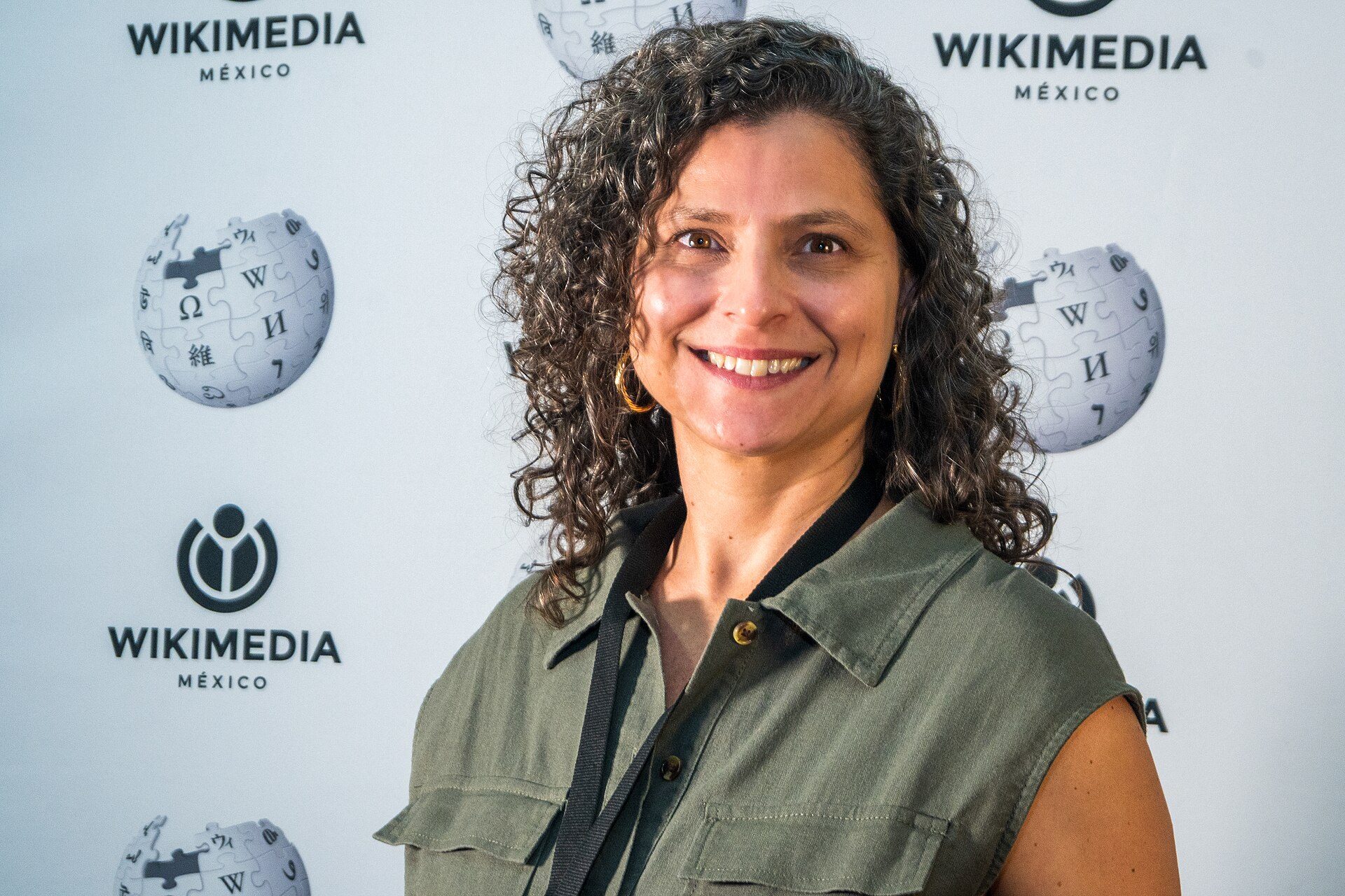A woman with curly hair smiling in front of a backdrop with logos.
