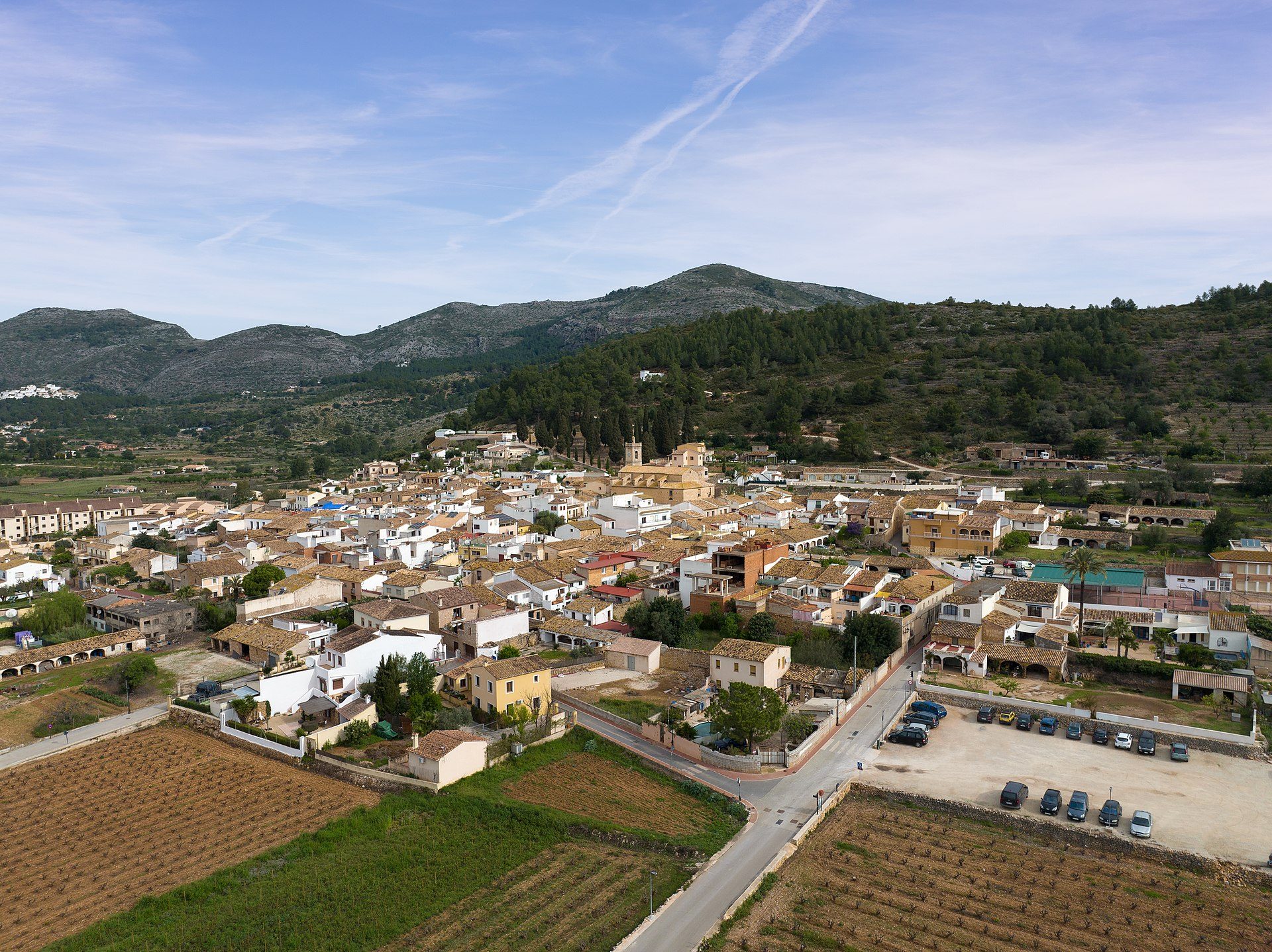 Aerial view of a quaint village with terracotta roofs, surrounded by lush green hills and a clear blue sky.