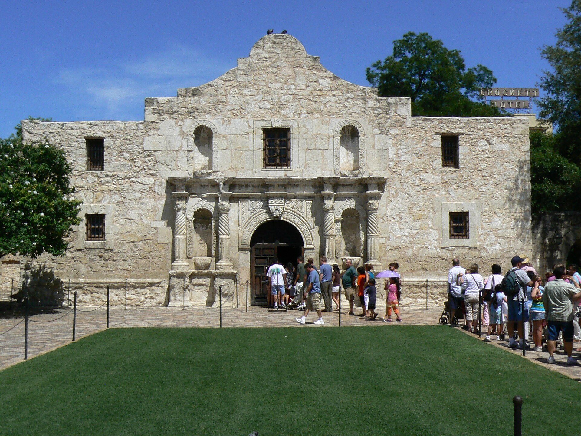 Alt text: Historic stone building with arched entrance, tourists gathered, green lawn in front.