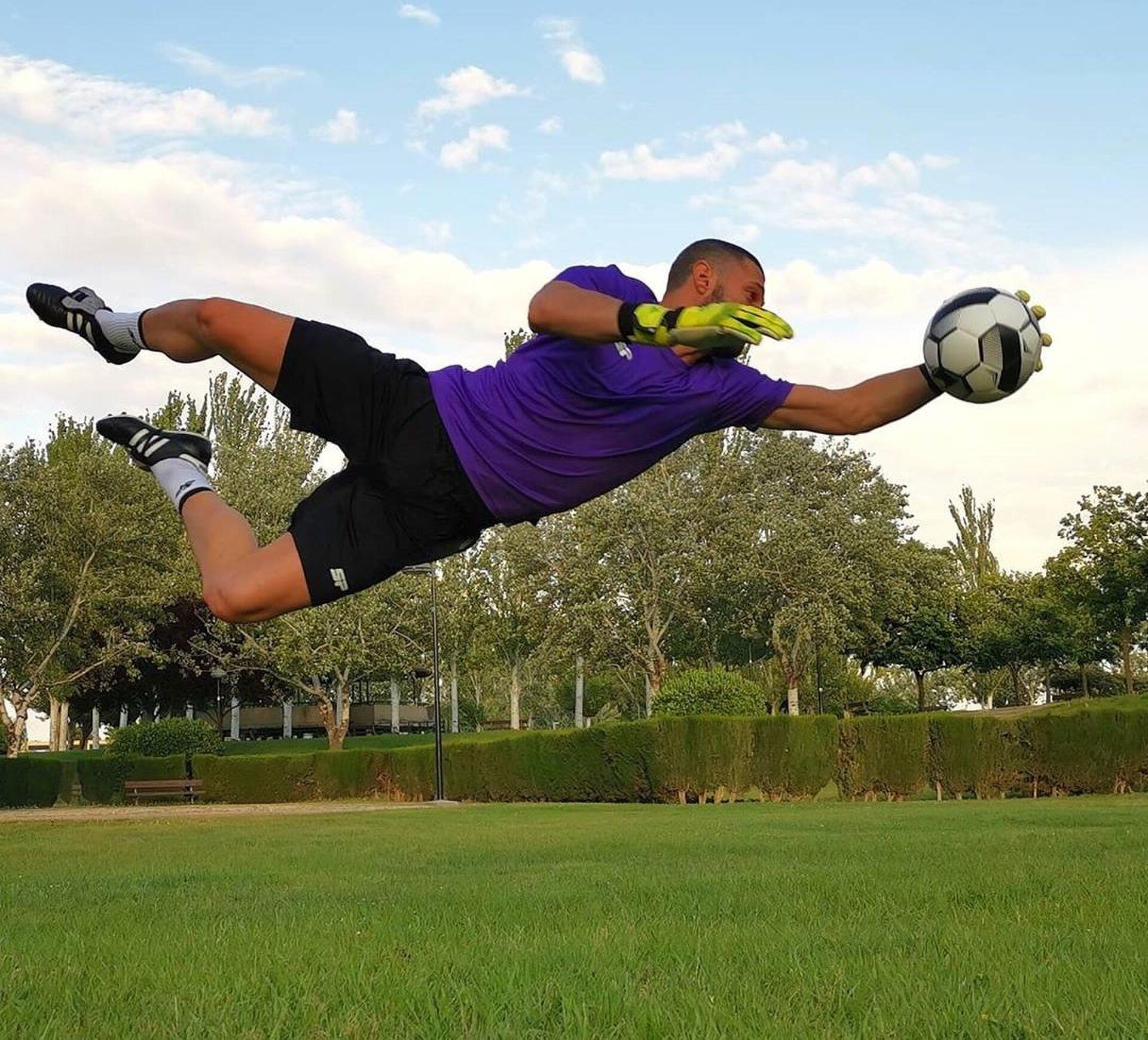 A soccer player in mid-air, wearing a purple shirt and black shorts, attempting to catch a soccer ball in a park.