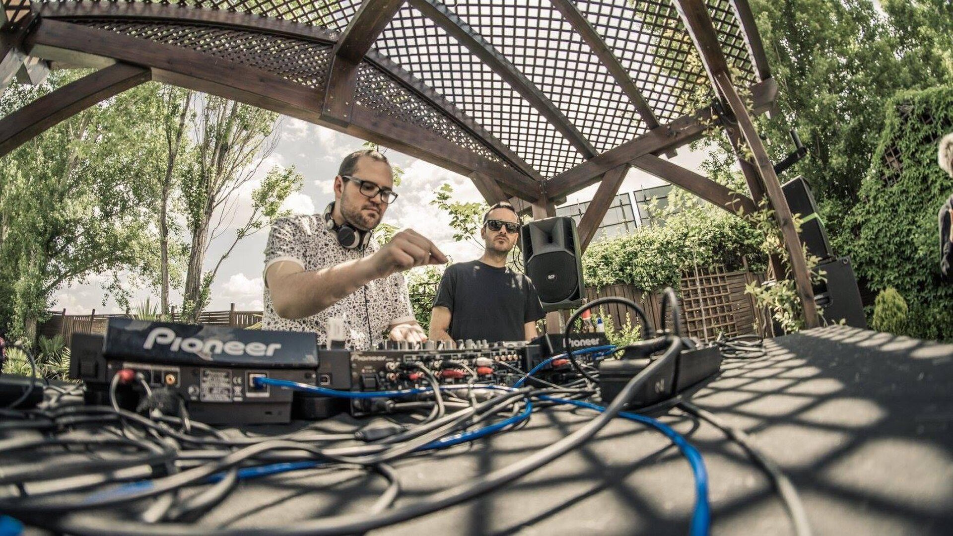 Two men operating DJ equipment under a wooden pergola with greenery in the background.