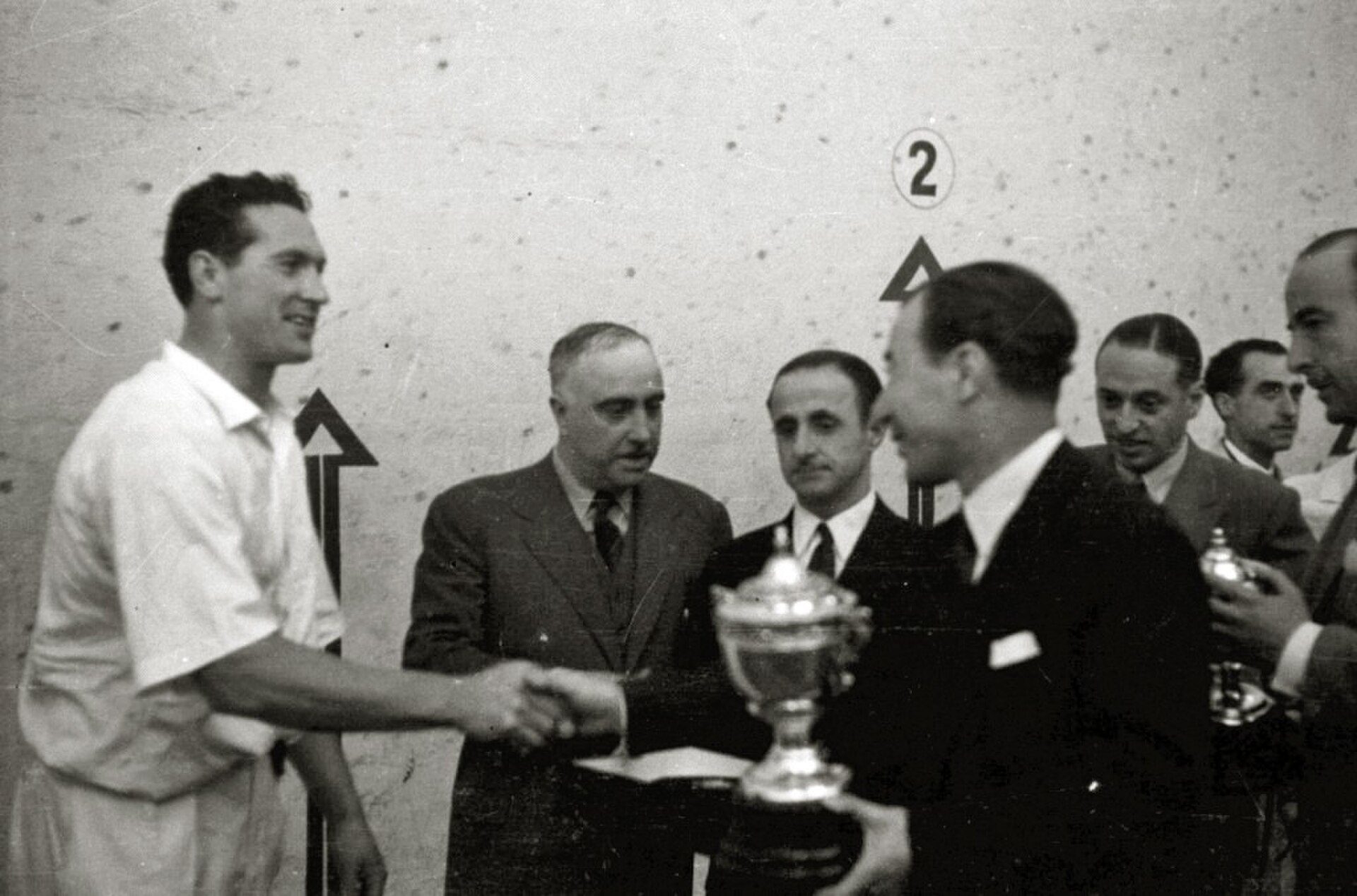 A black and white photo of a group of men in a room, one shaking hands with another holding a trophy.
