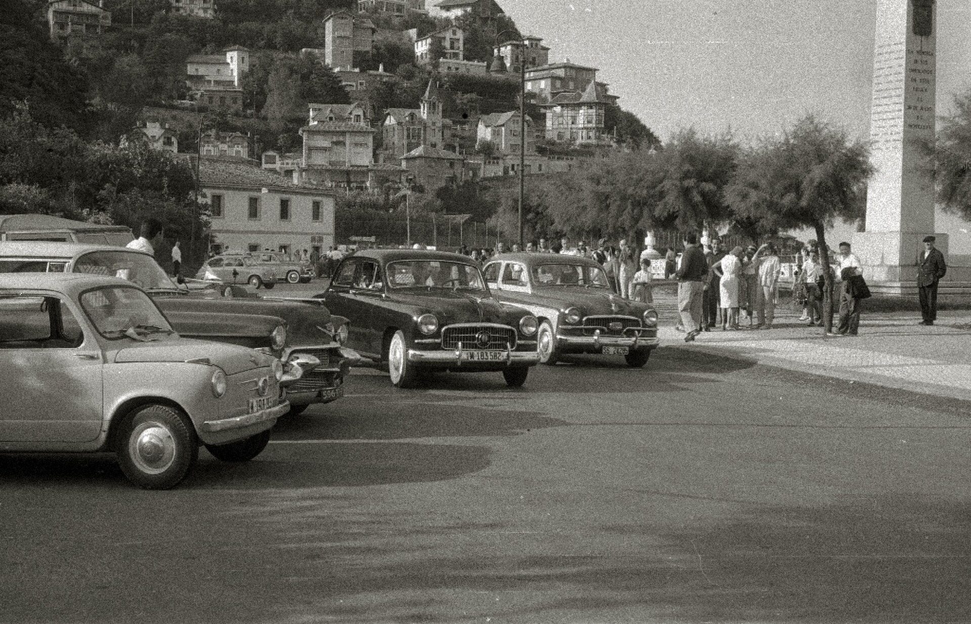 Alt text: Vintage street scene with classic cars, pedestrians, and a hillside town backdrop.