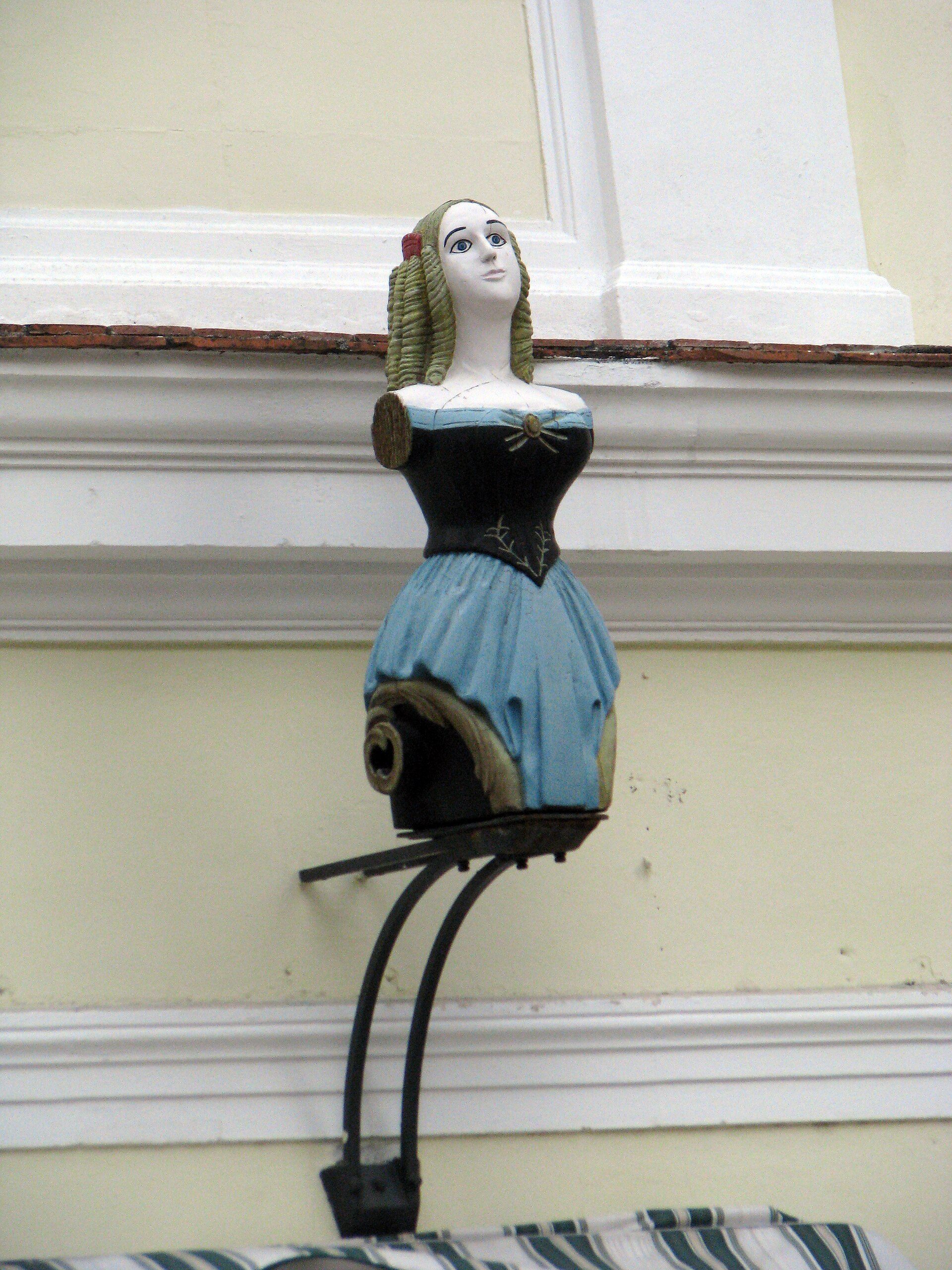 A vintage doll in a blue dress on a balcony with a view of a building.