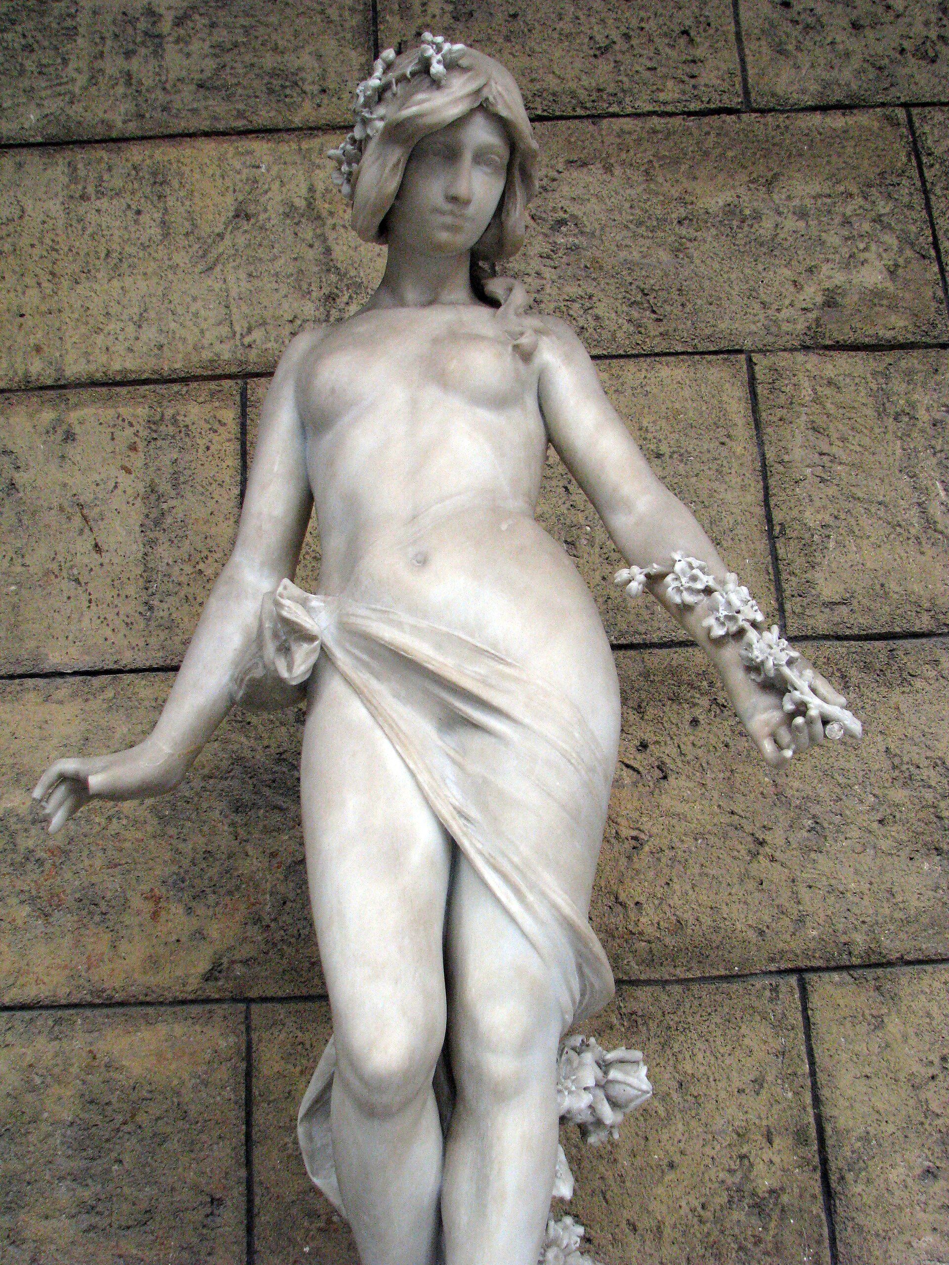 Alt text: A marble statue of a draped female figure with floral elements, set against a stone wall.