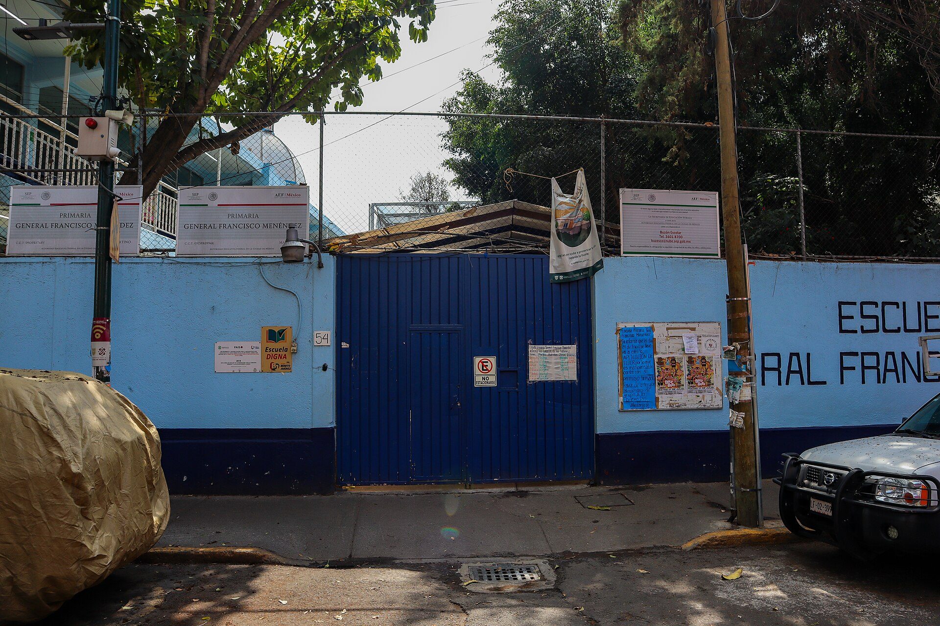 A blue gate with signs and posters, surrounded by trees and a parked car.