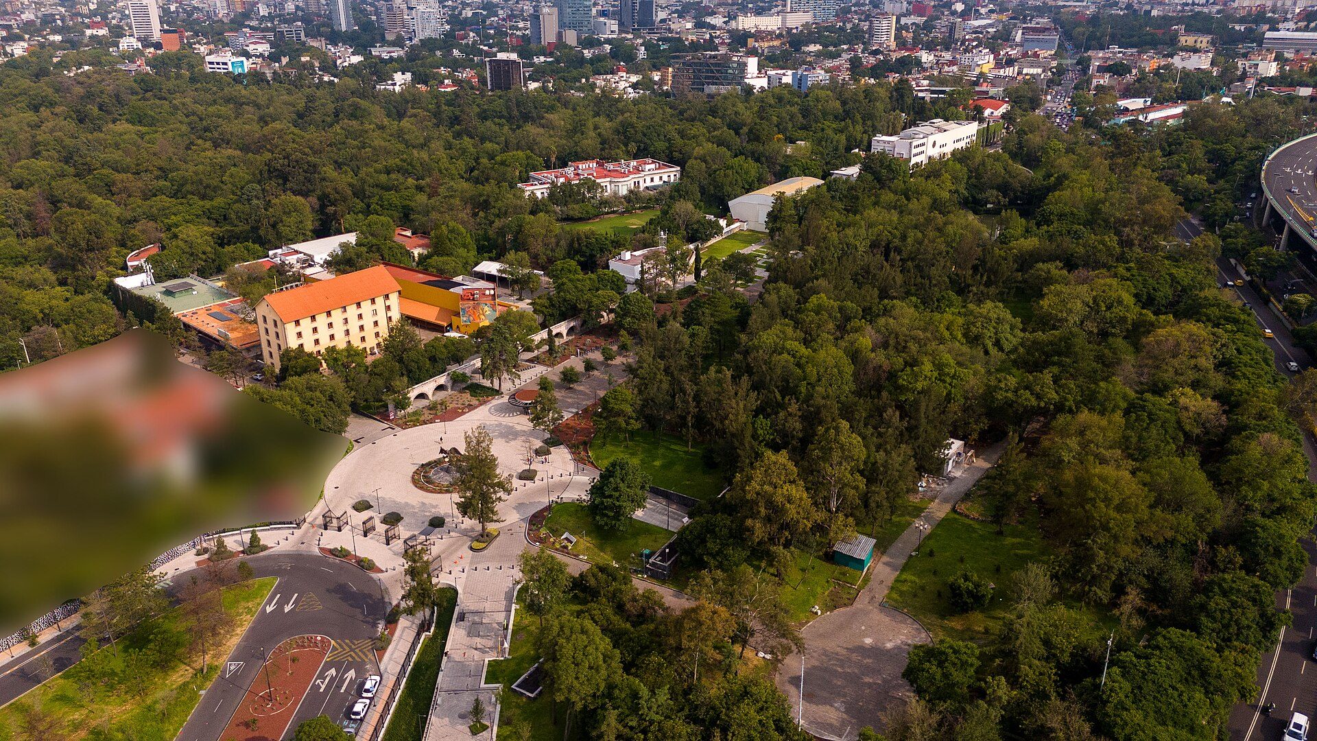 Aerial view of a hotel with a garden, pool, and cityscape in the background.
