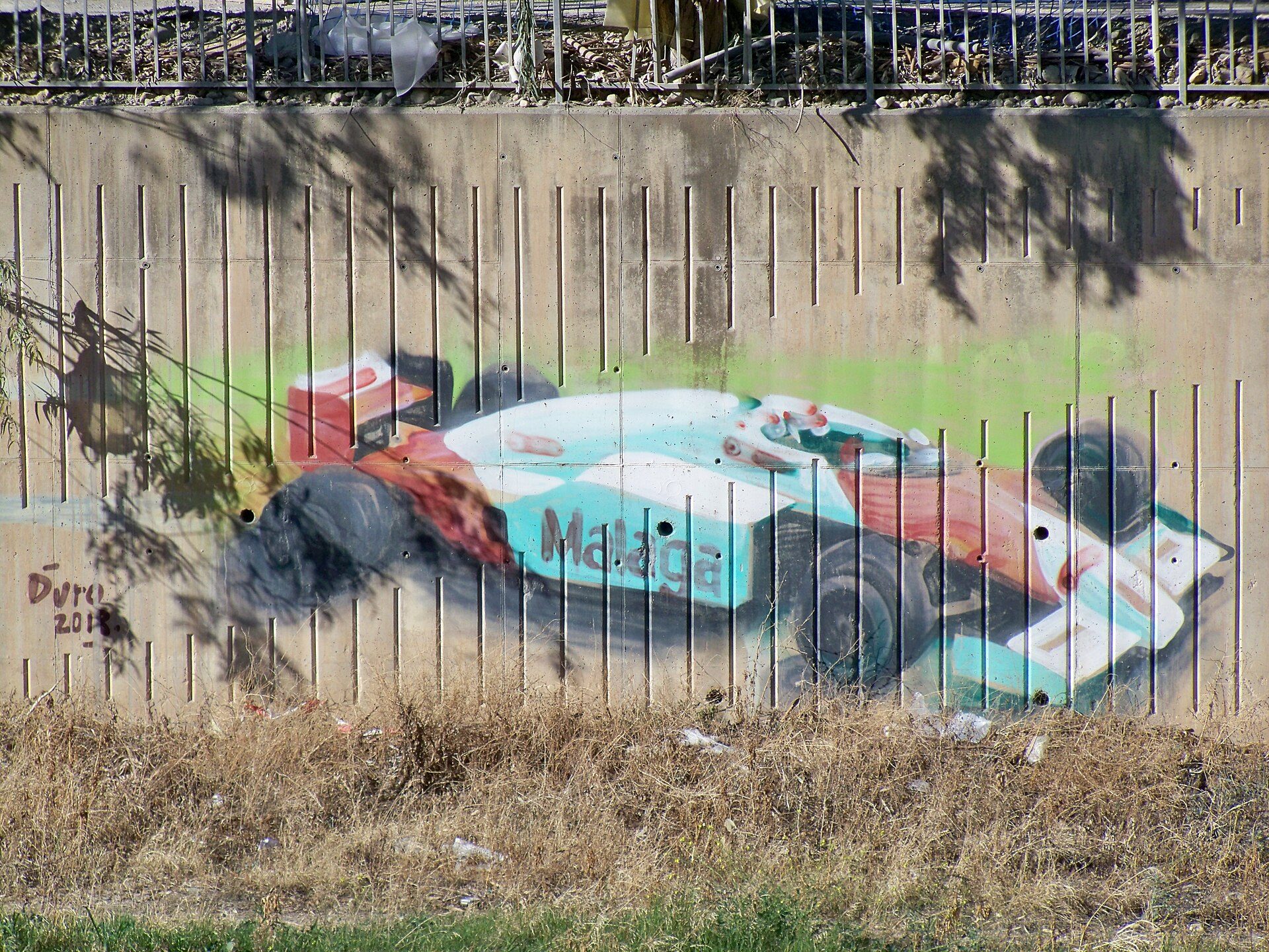A mural of a vintage race car on a concrete wall with a metal fence in front.