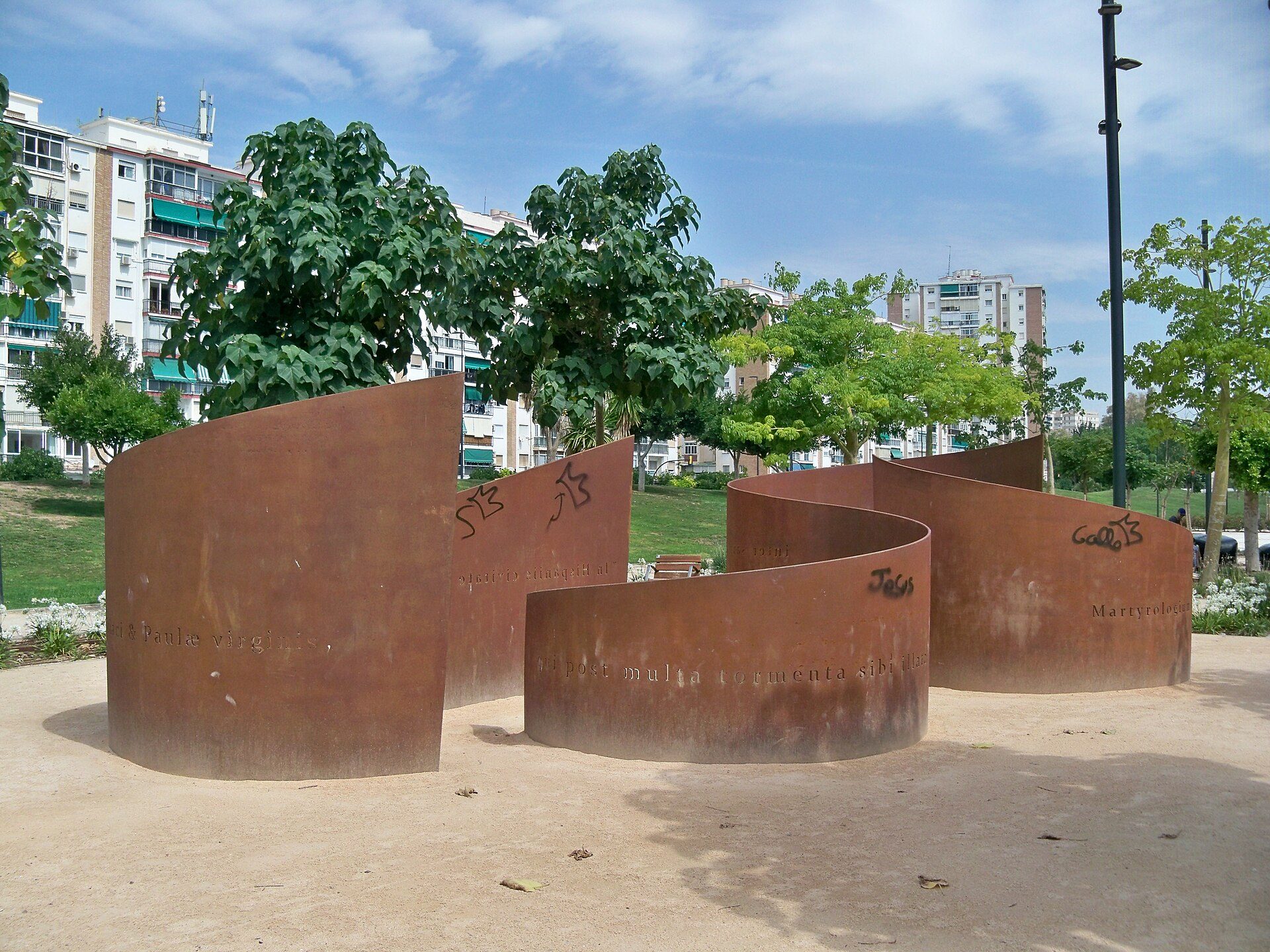 Outdoor sculpture installation with abstract shapes and inscriptions, surrounded by greenery and urban buildings.