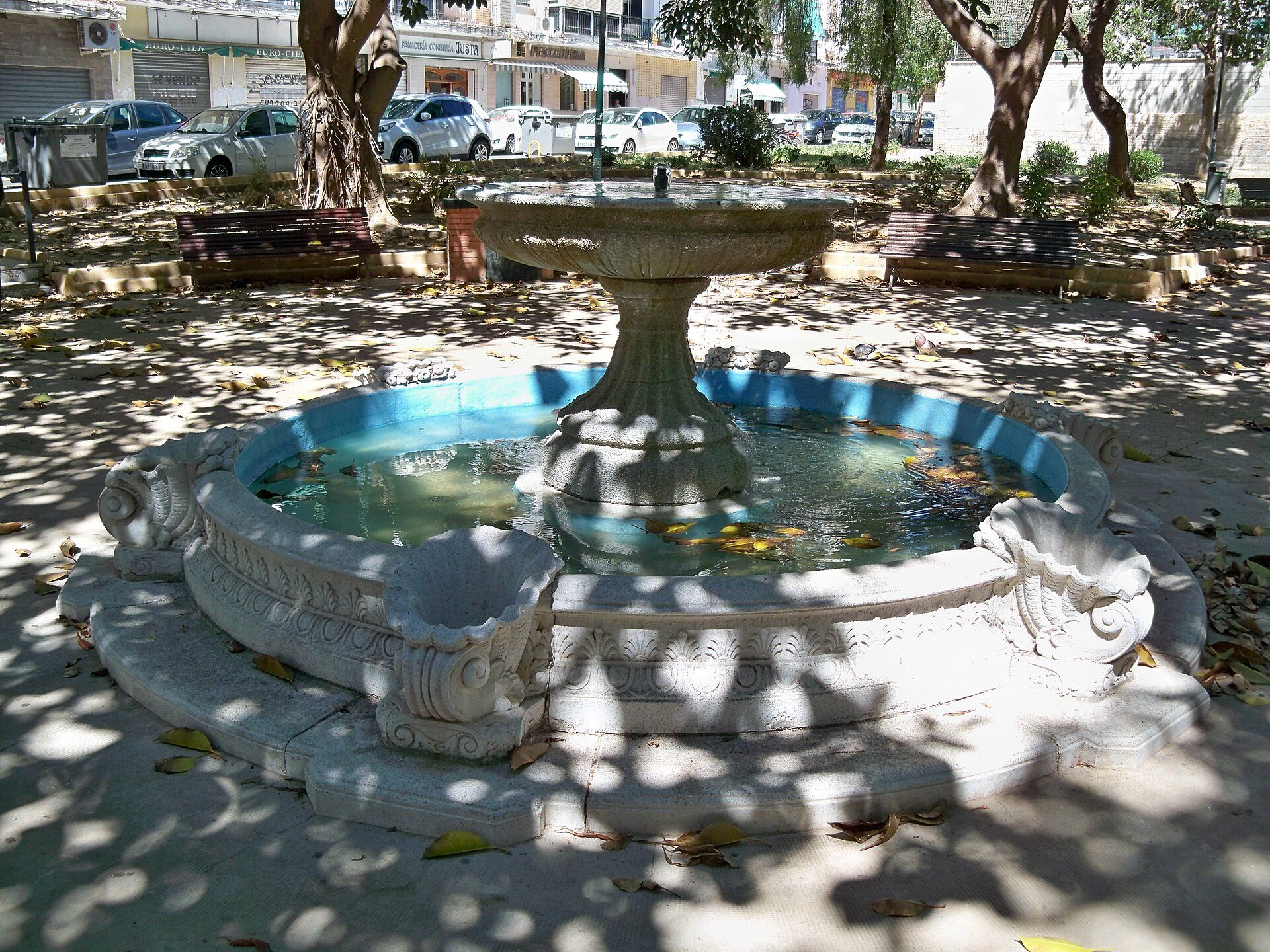 Alt text: Outdoor stone fountain with water, surrounded by benches and trees in a park setting.