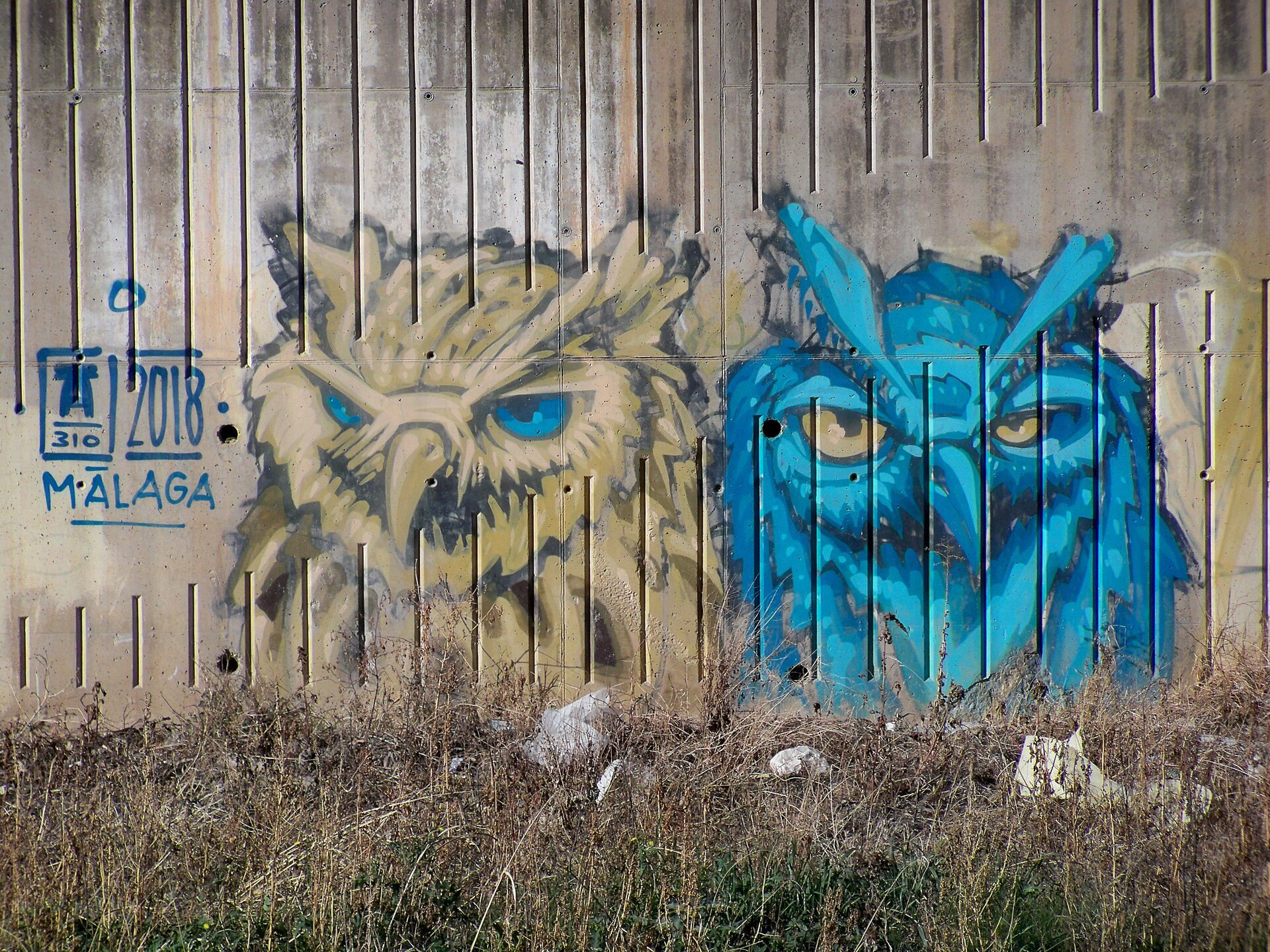 Two owl murals on a metal gate, one yellow, one blue, with "2018 MALAGA" text.