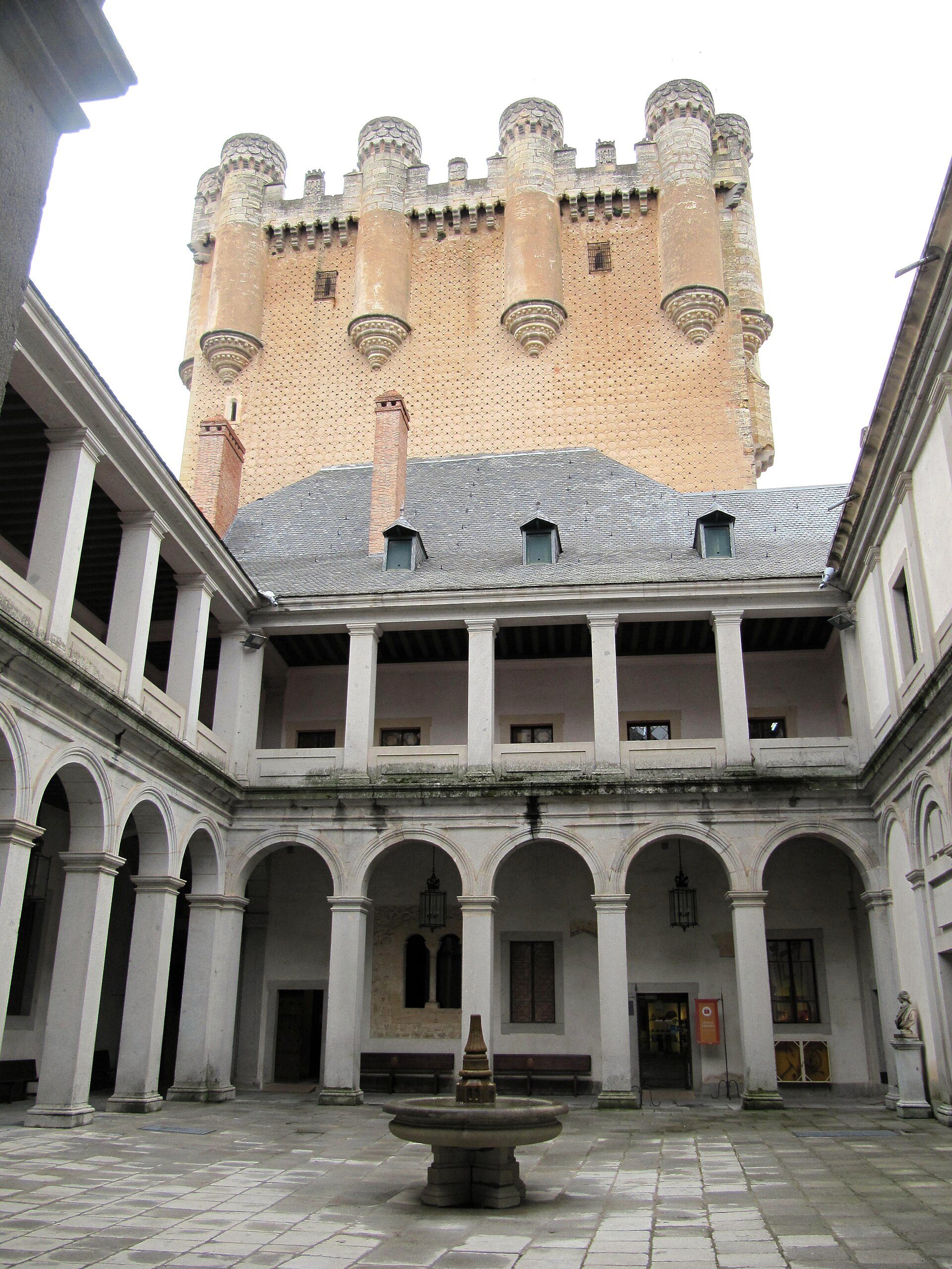 Courtyard with a central fountain, surrounded by arches and a tall tower in the background.