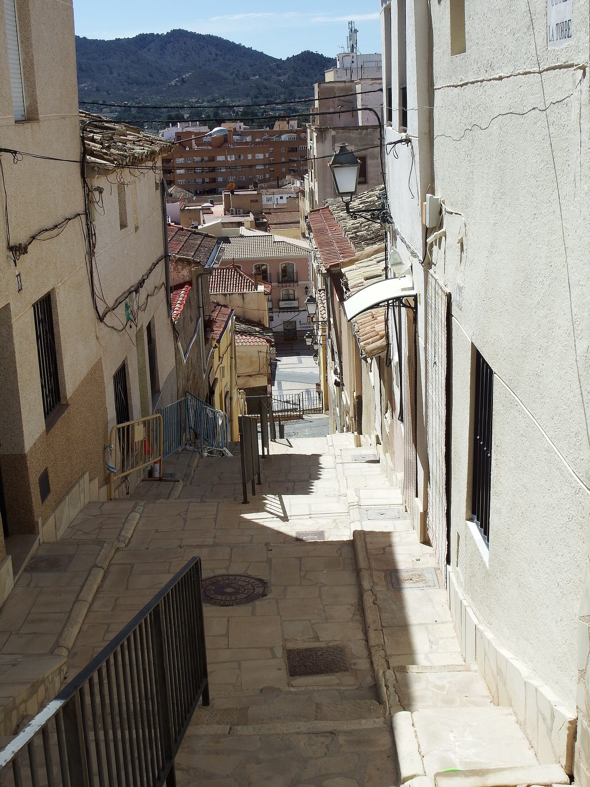 Narrow alley with stone pavement, leading to a mountain view, featuring old buildings and a street lamp.