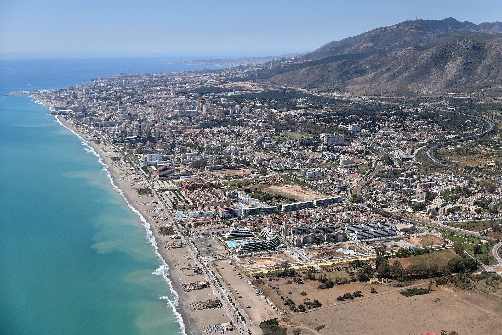 Aerial view of a coastal city with a beachfront, modern buildings, and a mountainous backdrop.