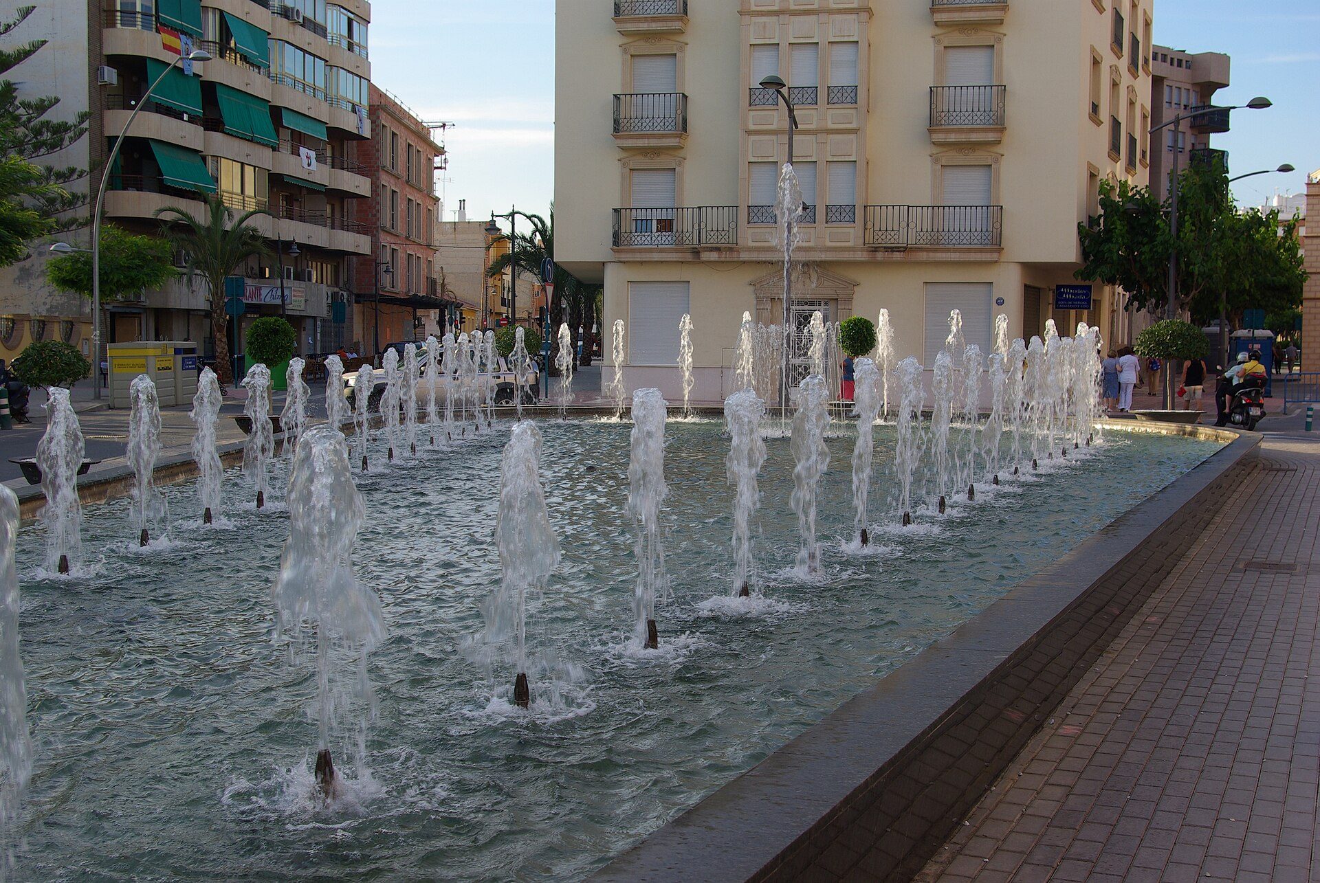 Alt text: Outdoor fountain with multiple jets, surrounded by urban buildings and palm trees.