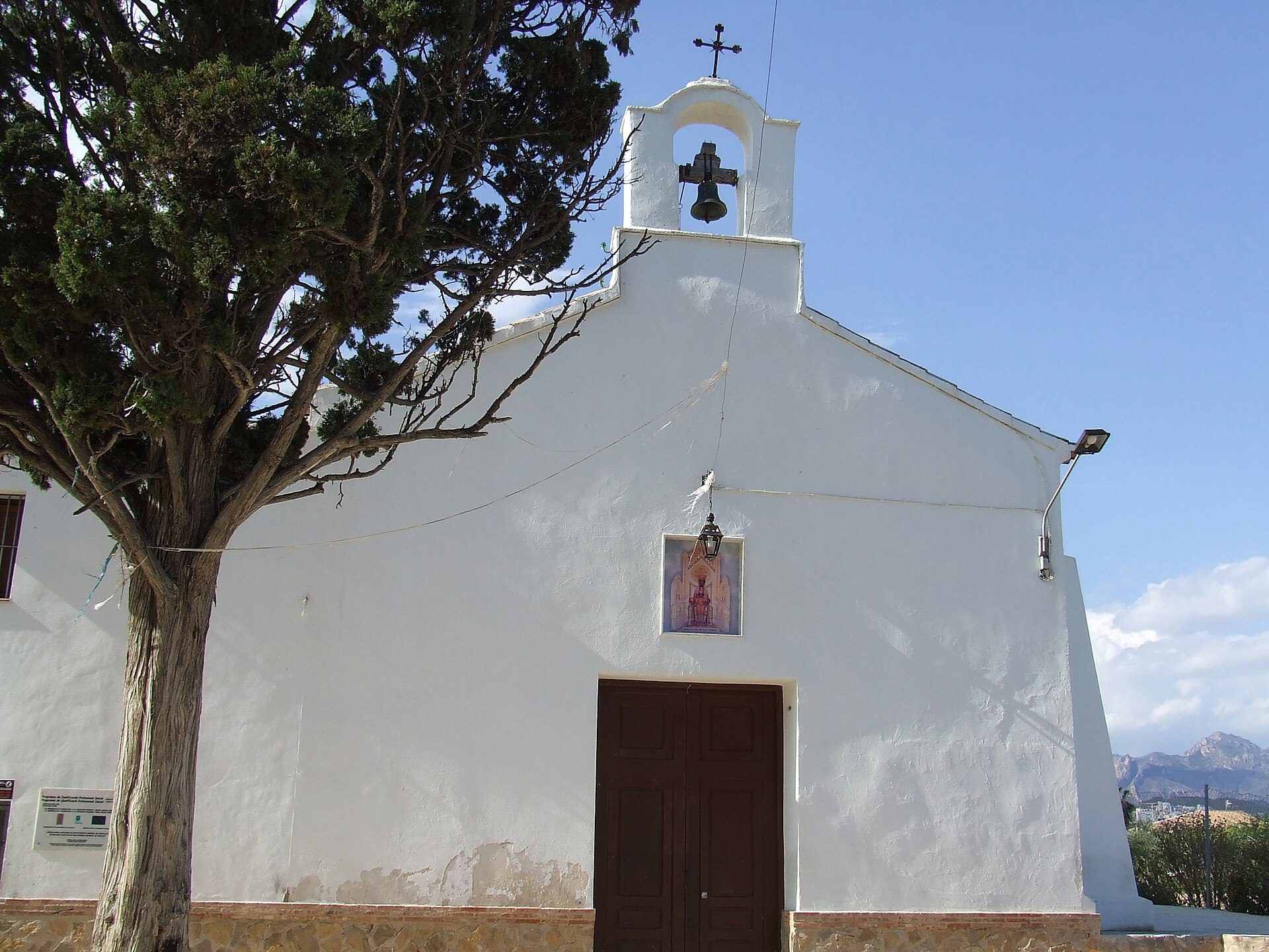 White-walled church with bell tower, tree, and clear blue sky.