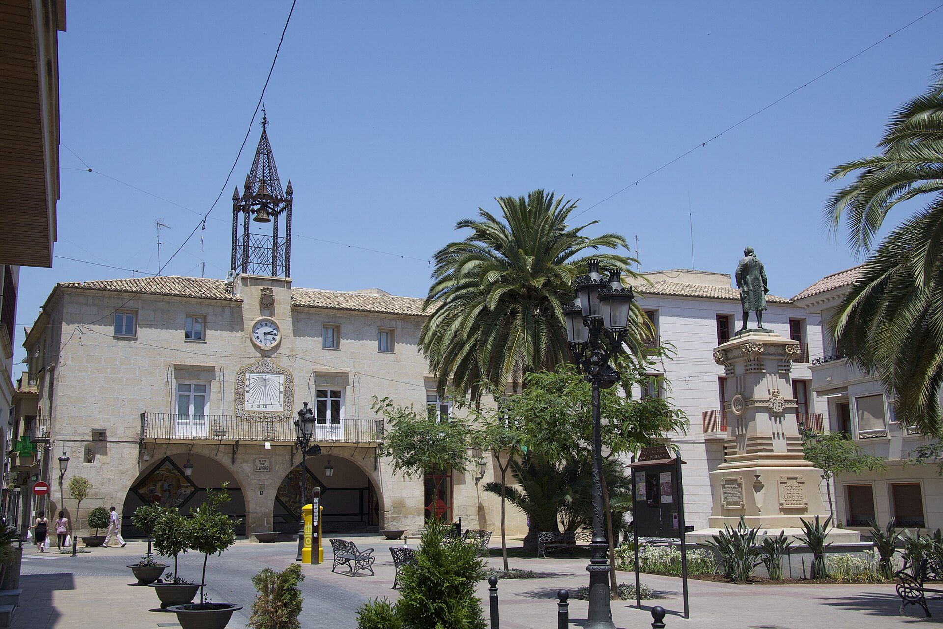 Alt text: Stone building with clock tower, palm trees, and statue in a sunny town square.
