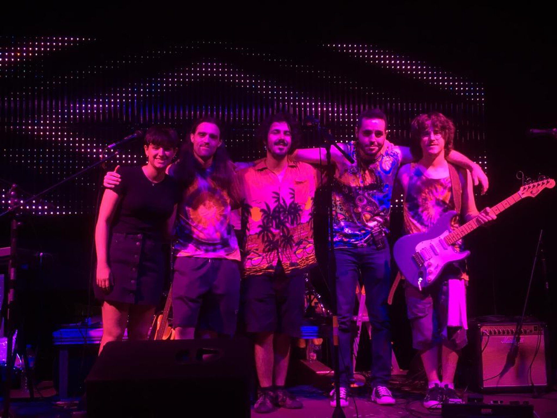 A group of five people posing on a stage with musical instruments and lighting.