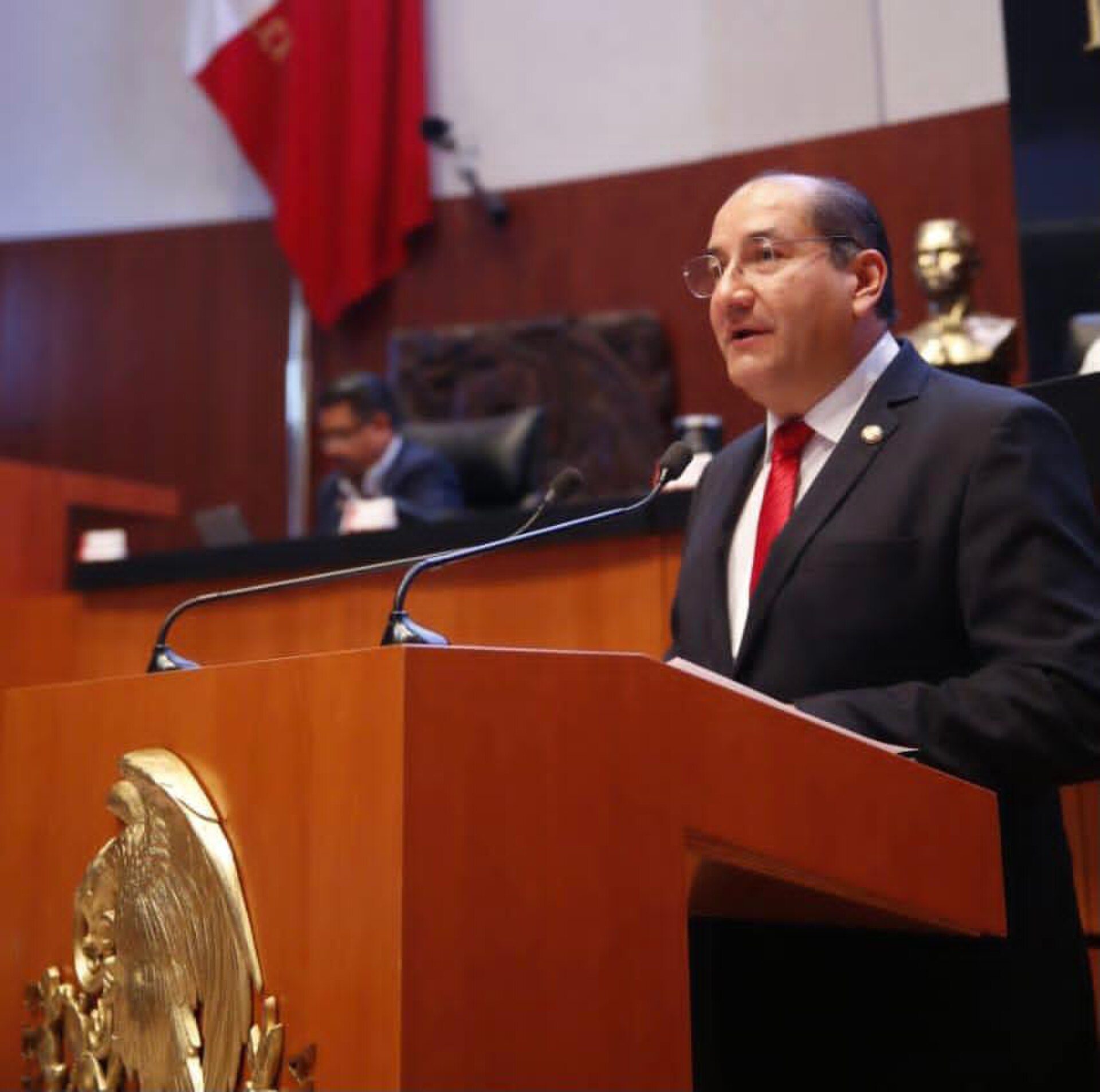 A man in a suit and red tie stands at a podium in a formal government chamber.