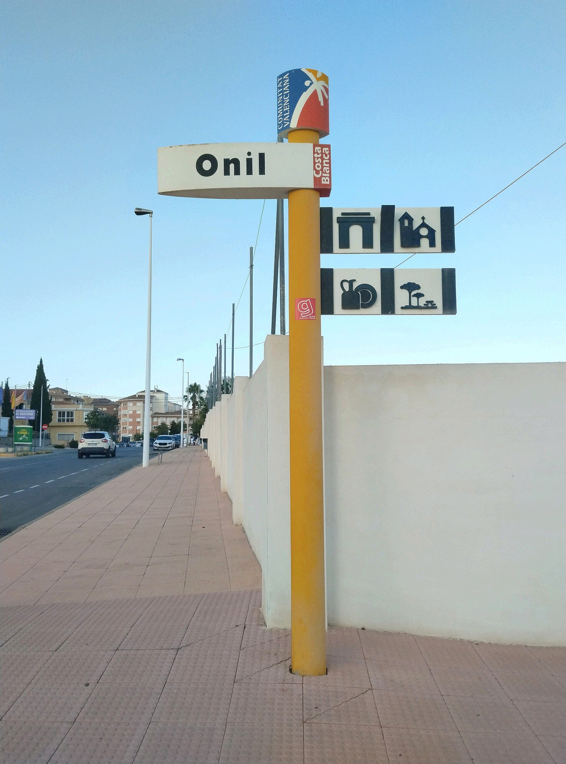 Alt text: Outdoor signpost with "Onil" and bus stop icons, near a road with buildings in the background.