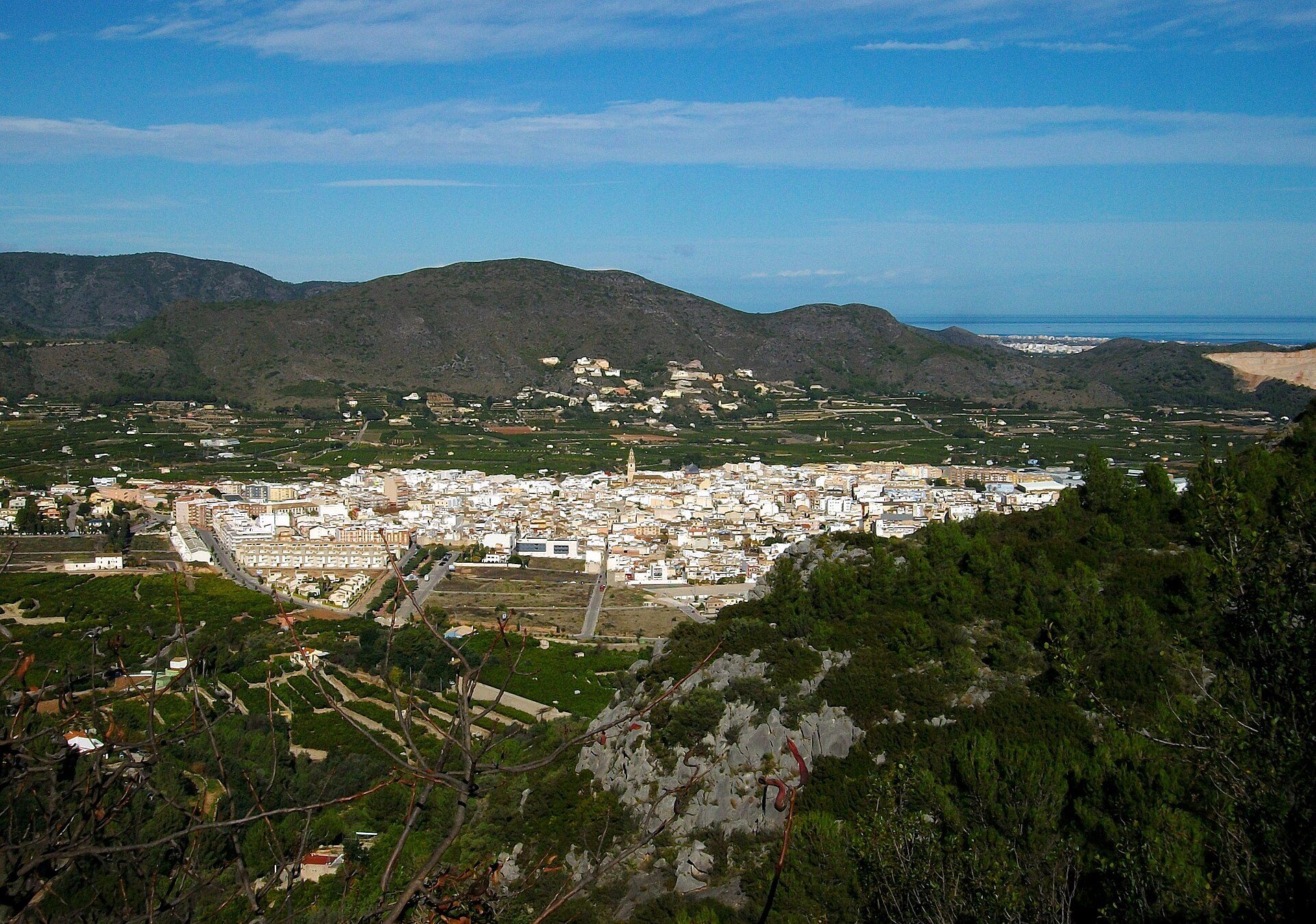 Alt text: Panoramic view of a coastal town with white buildings, greenery, and distant mountains.