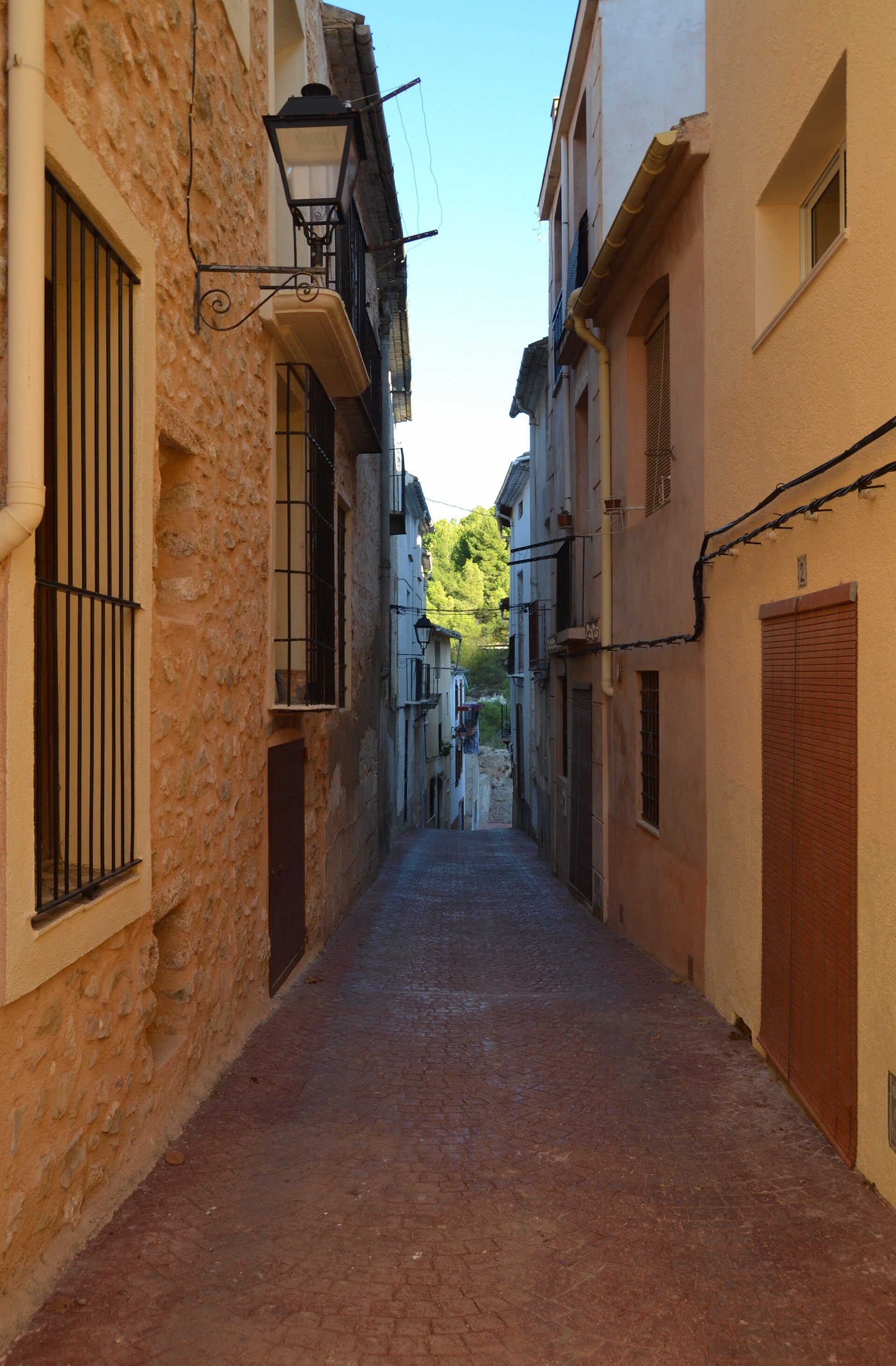 Narrow alley with stone walls, metal window grilles, and a view of greenery at the end.
