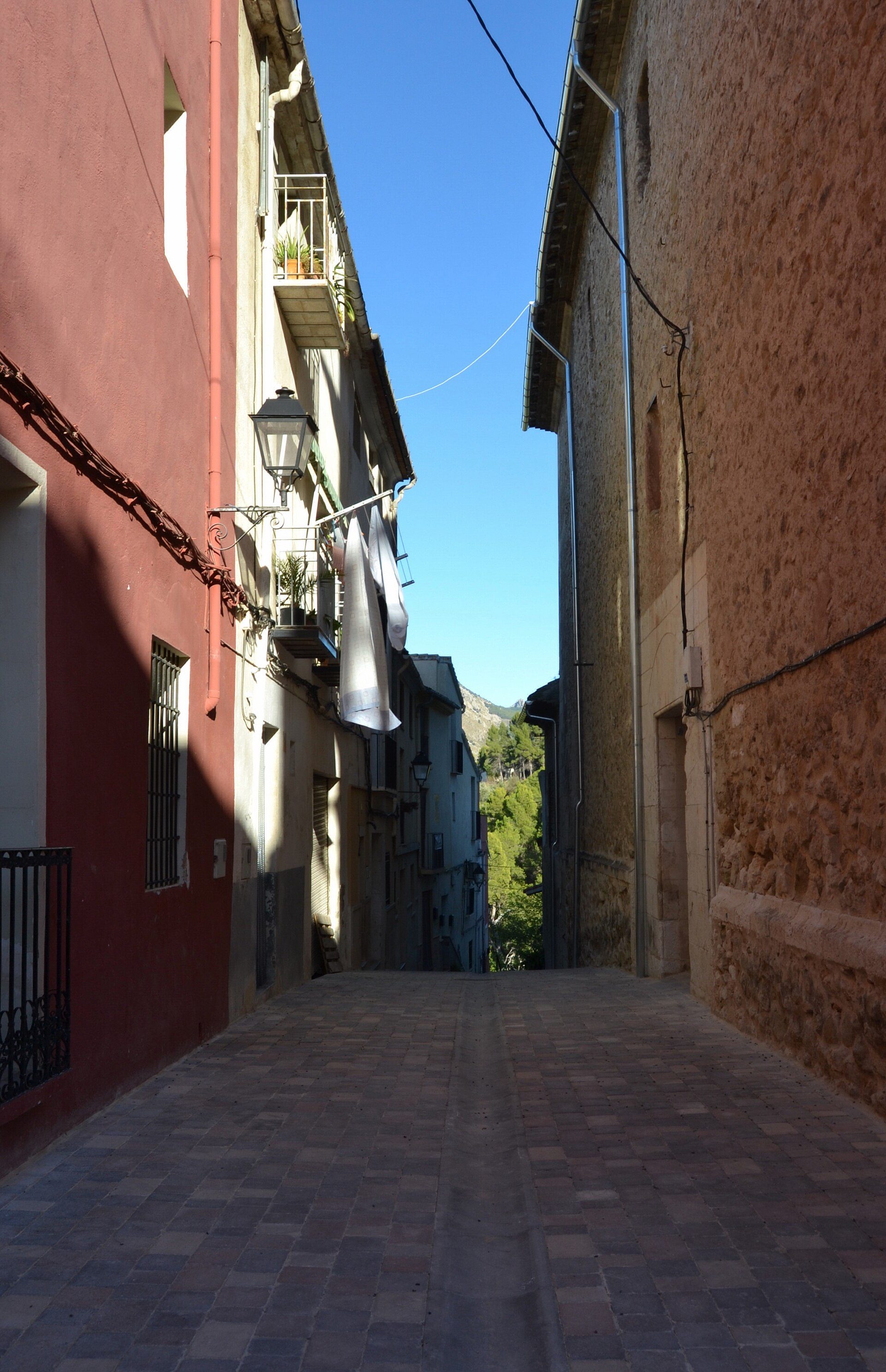 Narrow alley with red and beige buildings, clothes hanging, clear blue sky.