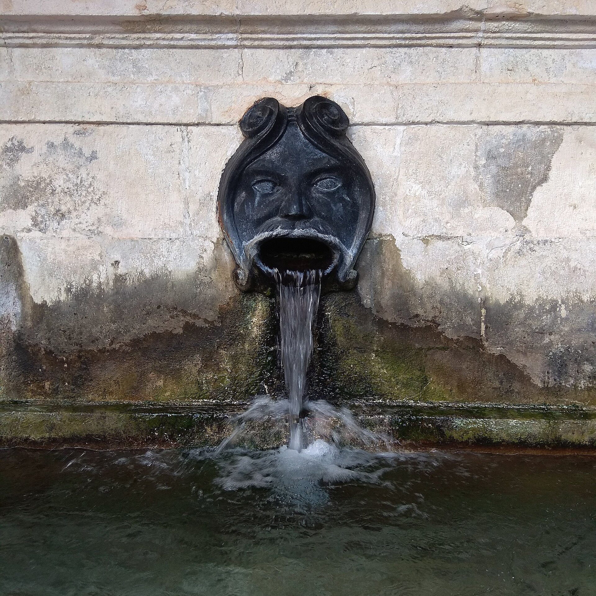 An ornate stone fountain with a face spout, water flowing into a basin.