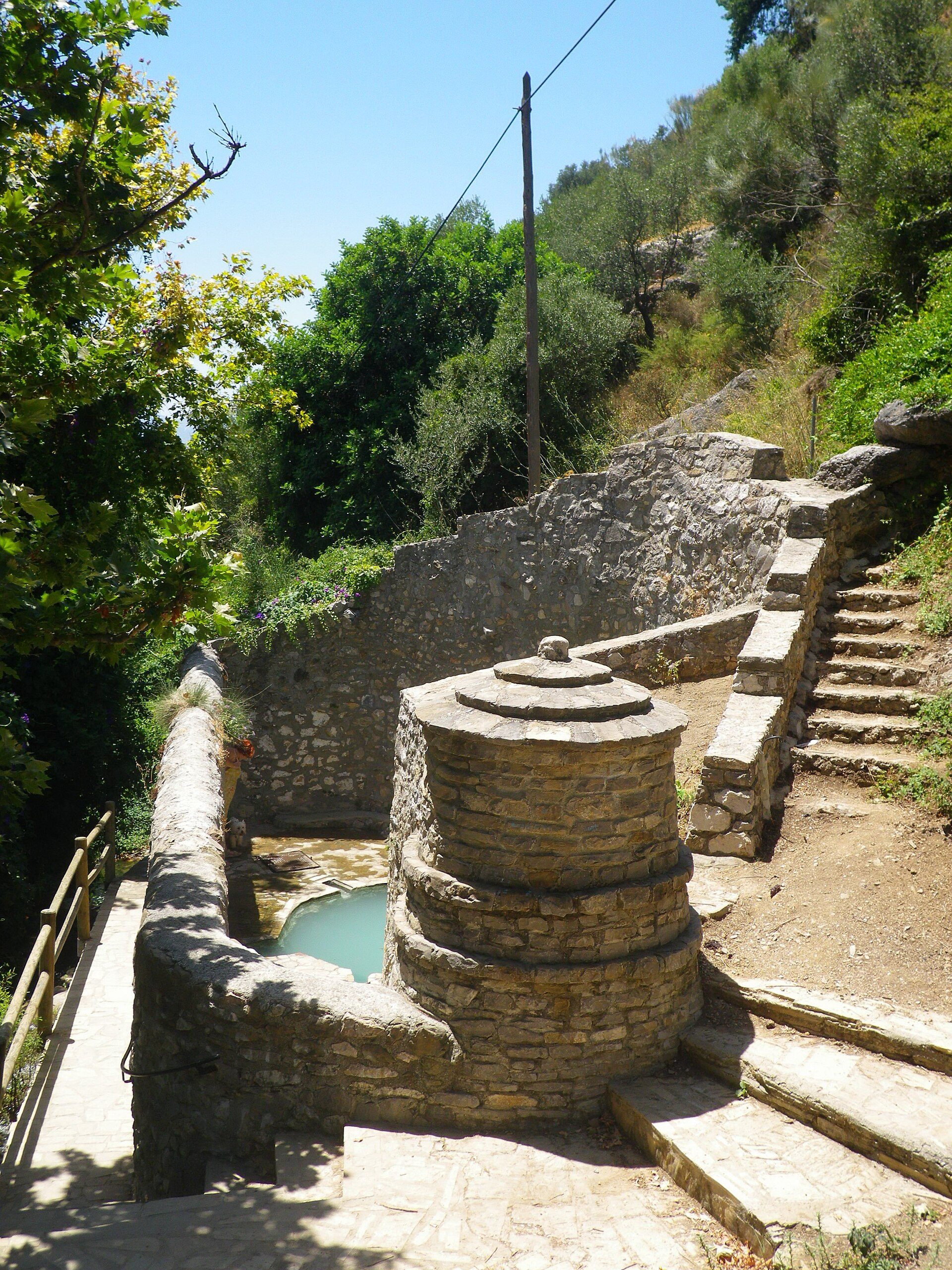 Alt text: Stone-built outdoor pool with steps, surrounded by greenery and a clear blue sky.