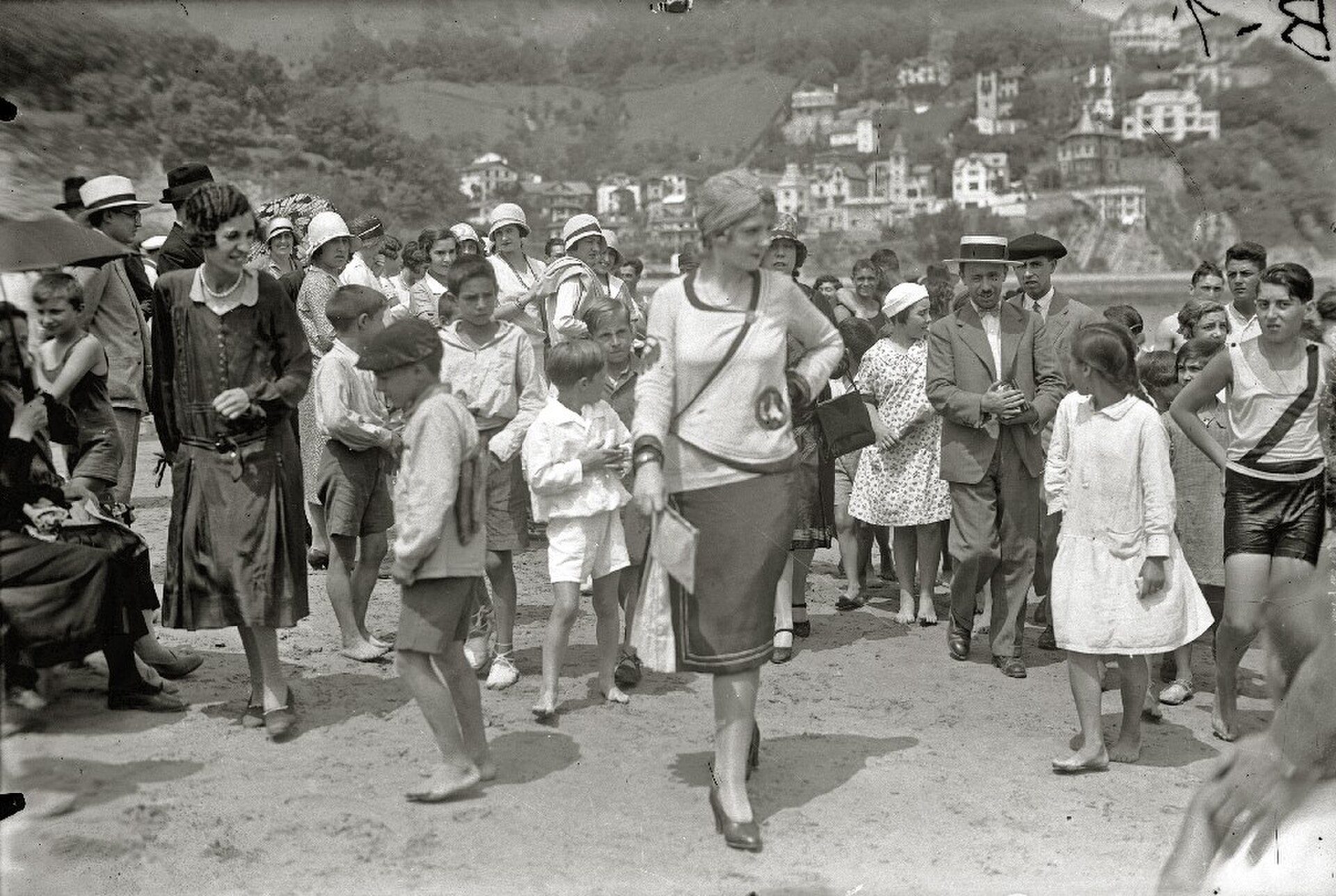 Alt text: Vintage beach scene with people walking, hills, and buildings in the background.