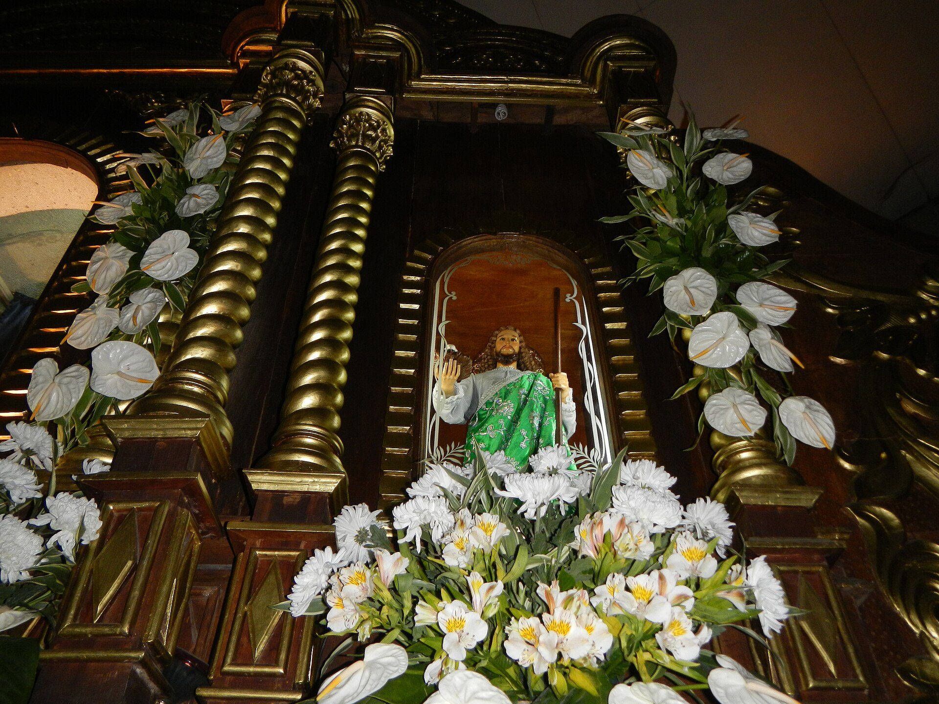 Alt text: Ornate religious statue in a gated alcove surrounded by white flowers and gold columns.