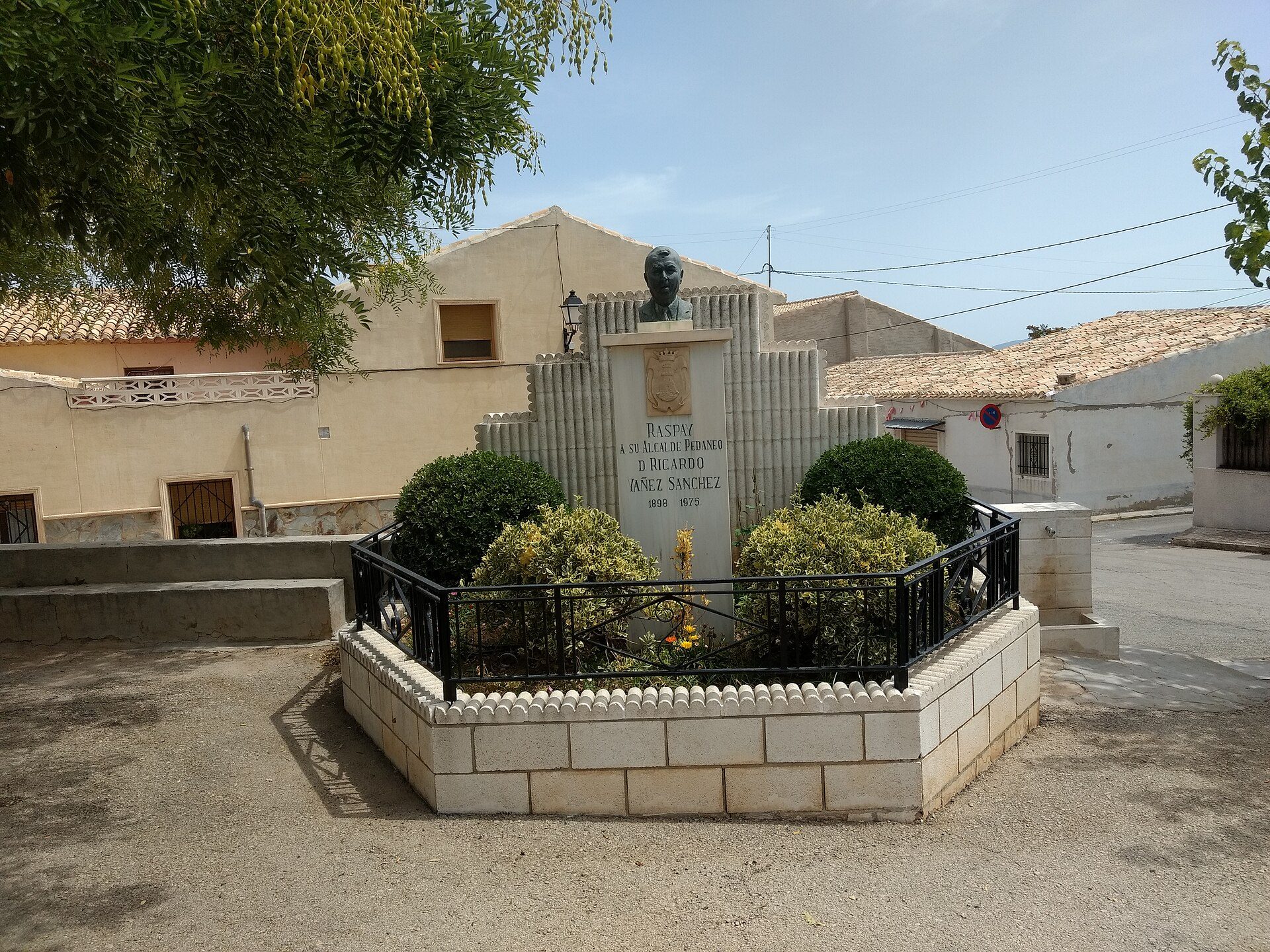 Alt text: Outdoor monument with bust, surrounded by greenery, in a courtyard with buildings in the background.