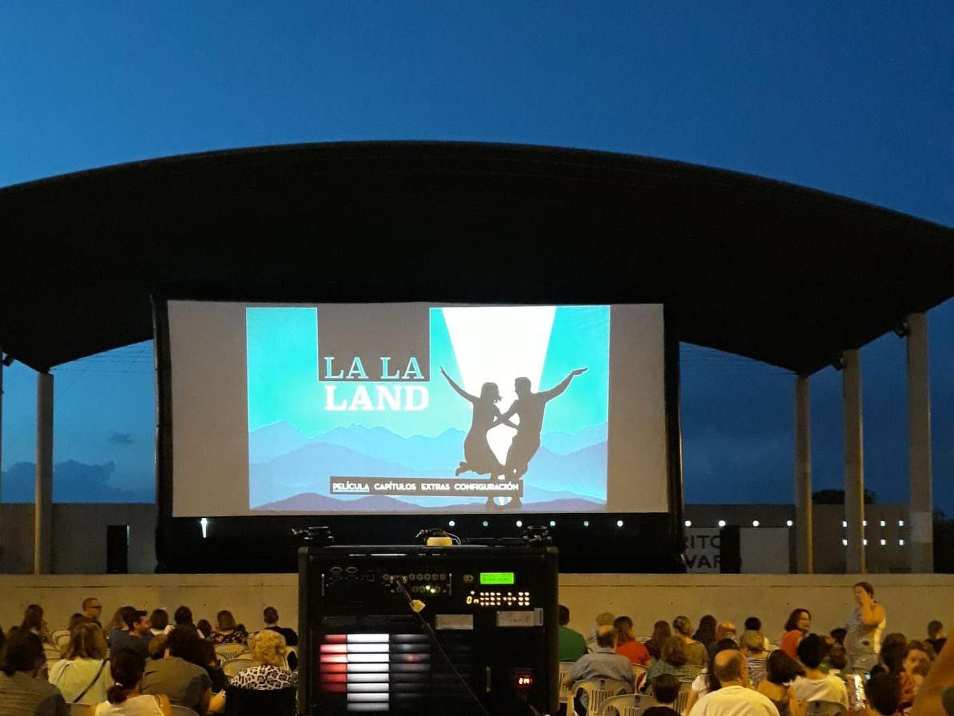 An outdoor event with a large screen displaying "LA LA LAND" and a crowd of people watching.