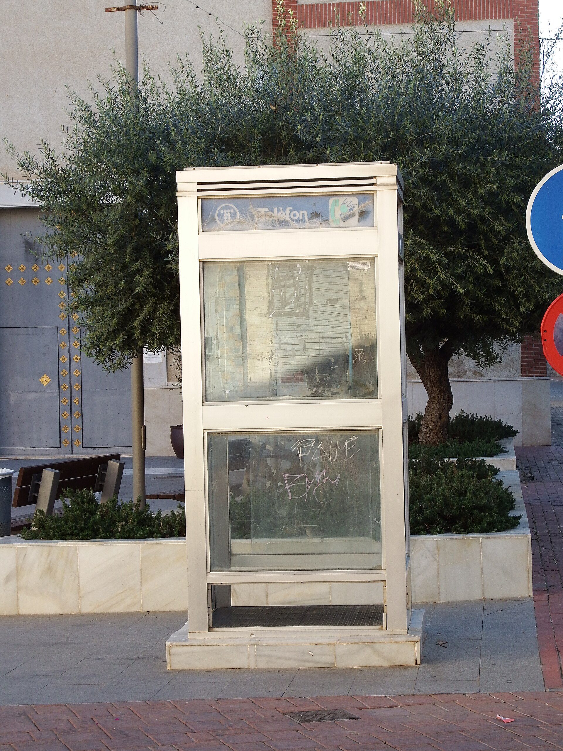 A public phone booth on a sidewalk with a tree and building in the background.
