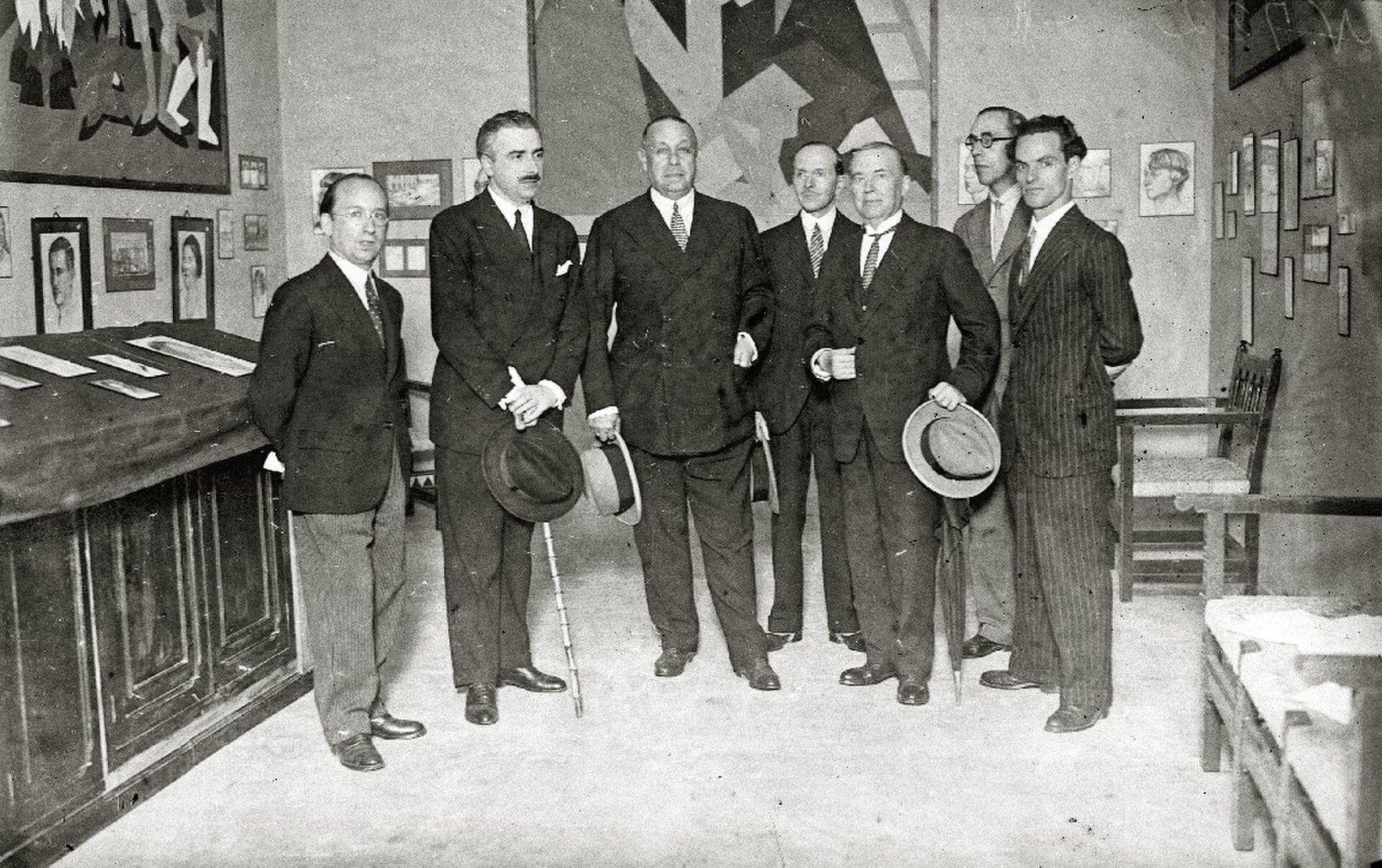A group of men in suits standing in an office with a large desk and framed pictures on the wall.
